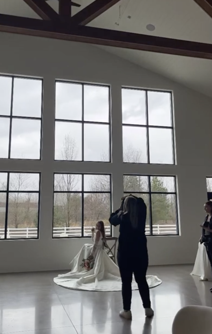 A bride sitting with a bouquet in front of large windows in a bright room, while a photographer takes her picture and a woman with a notepad stands nearby.