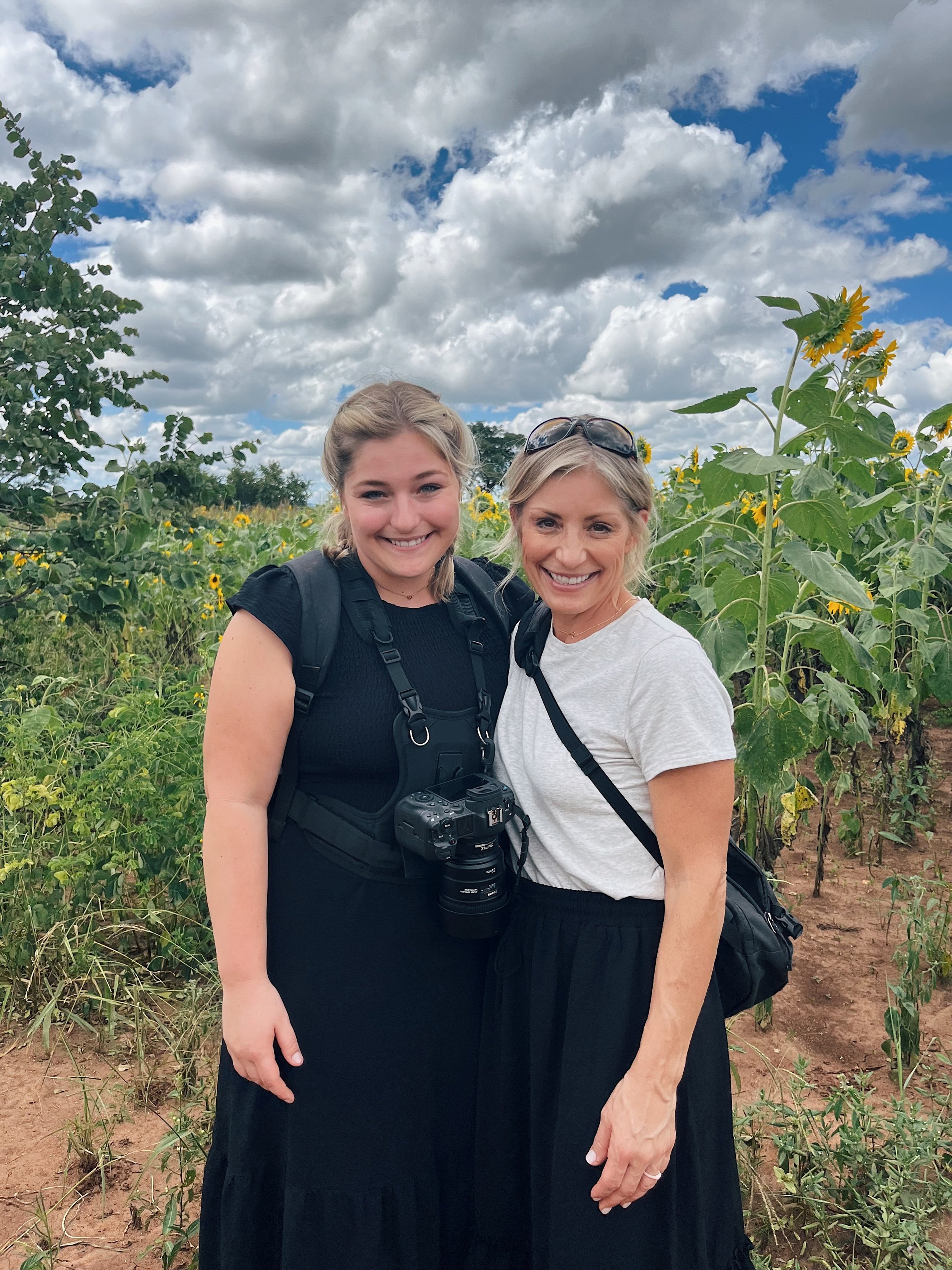 Two women smiling and standing close together in a field of sunflowers under a partly cloudy sky. One woman has blonde hair and sunglasses on her head, and the other has light brown hair. They are wearing casual clothing and backpacks.