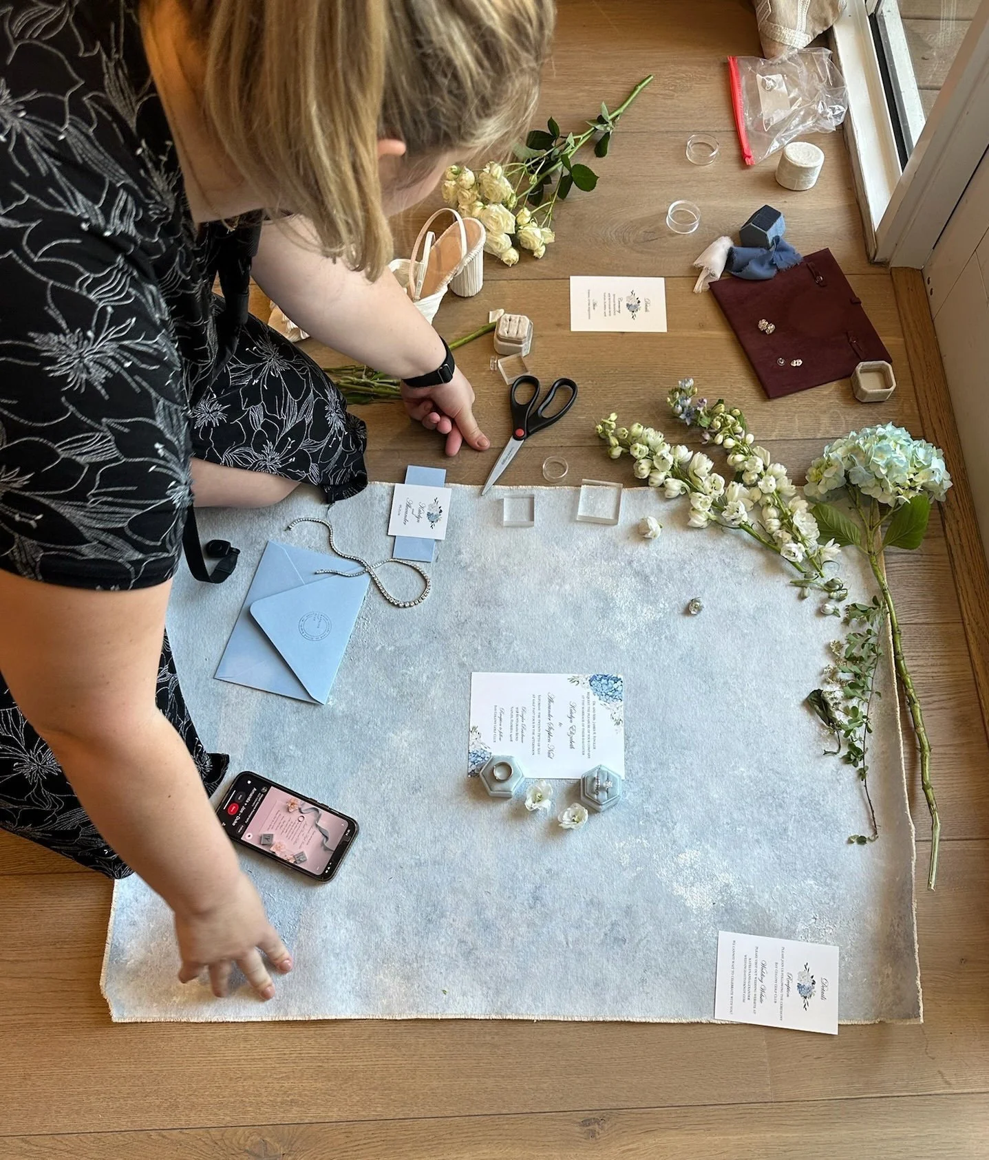 A person arranging wedding or event decorations including white flowers, small vases, a jewelry box, and decorative items on a table covered with a white cloth. The table also has a few printed papers and an iPhone.