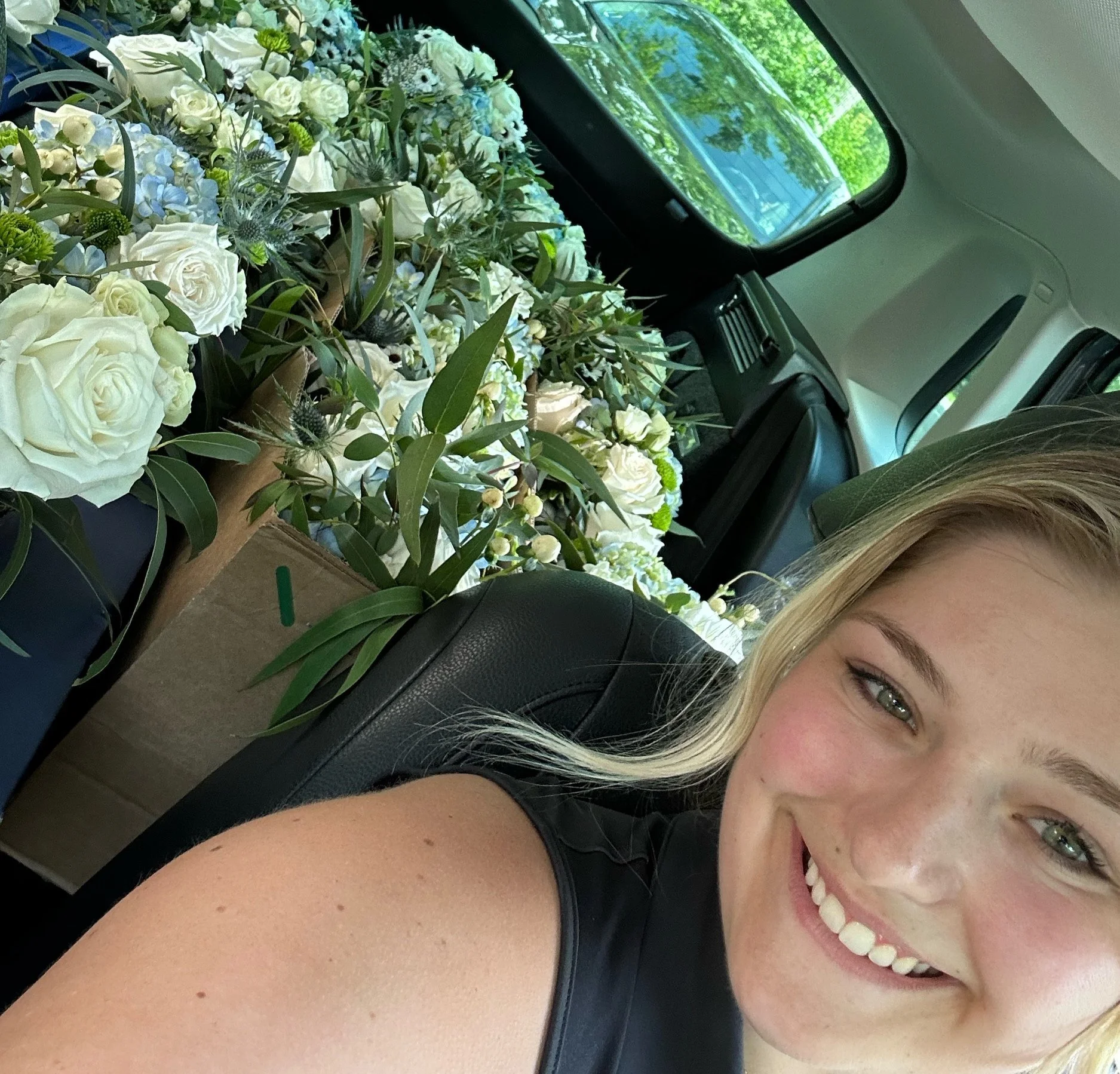 A smiling woman with blonde hair and glasses wearing a black sleeveless top, sitting in a car. The woman is close to the camera on the right side, showing part of her face and arm. The background inside the car features a table full of white and ligh