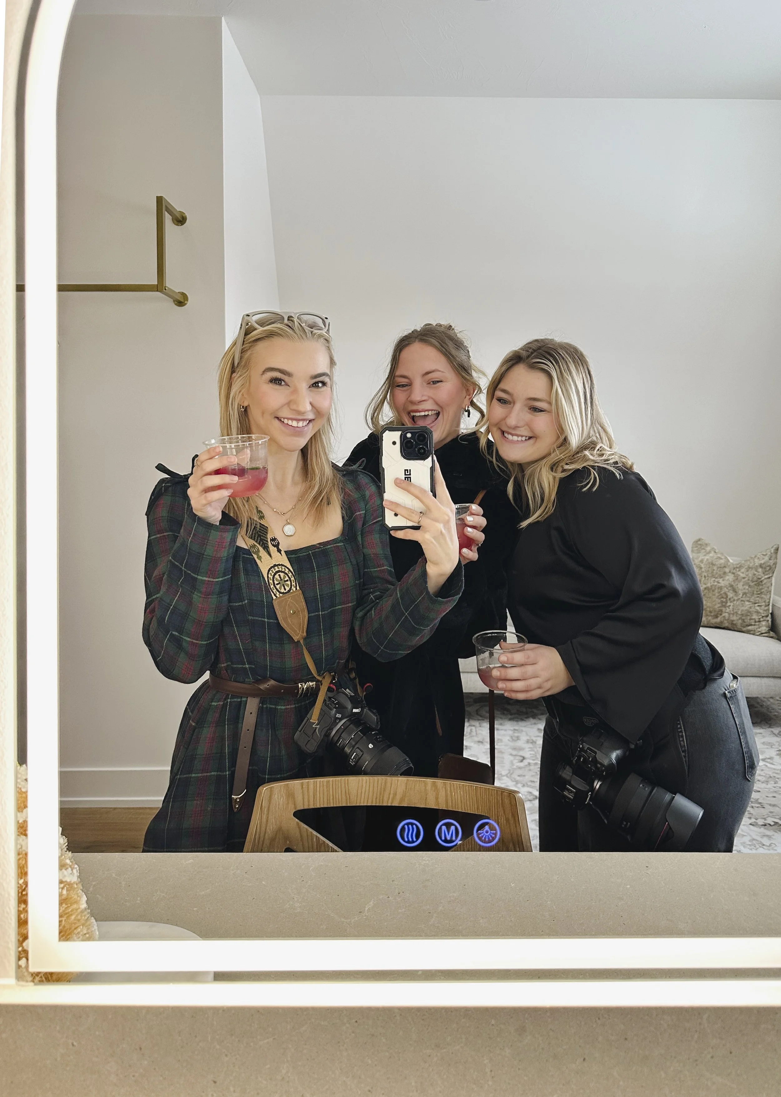 Three women taking a selfie in front of a mirror, all smiling and holding drinks. Two women have cameras hanging around their necks.