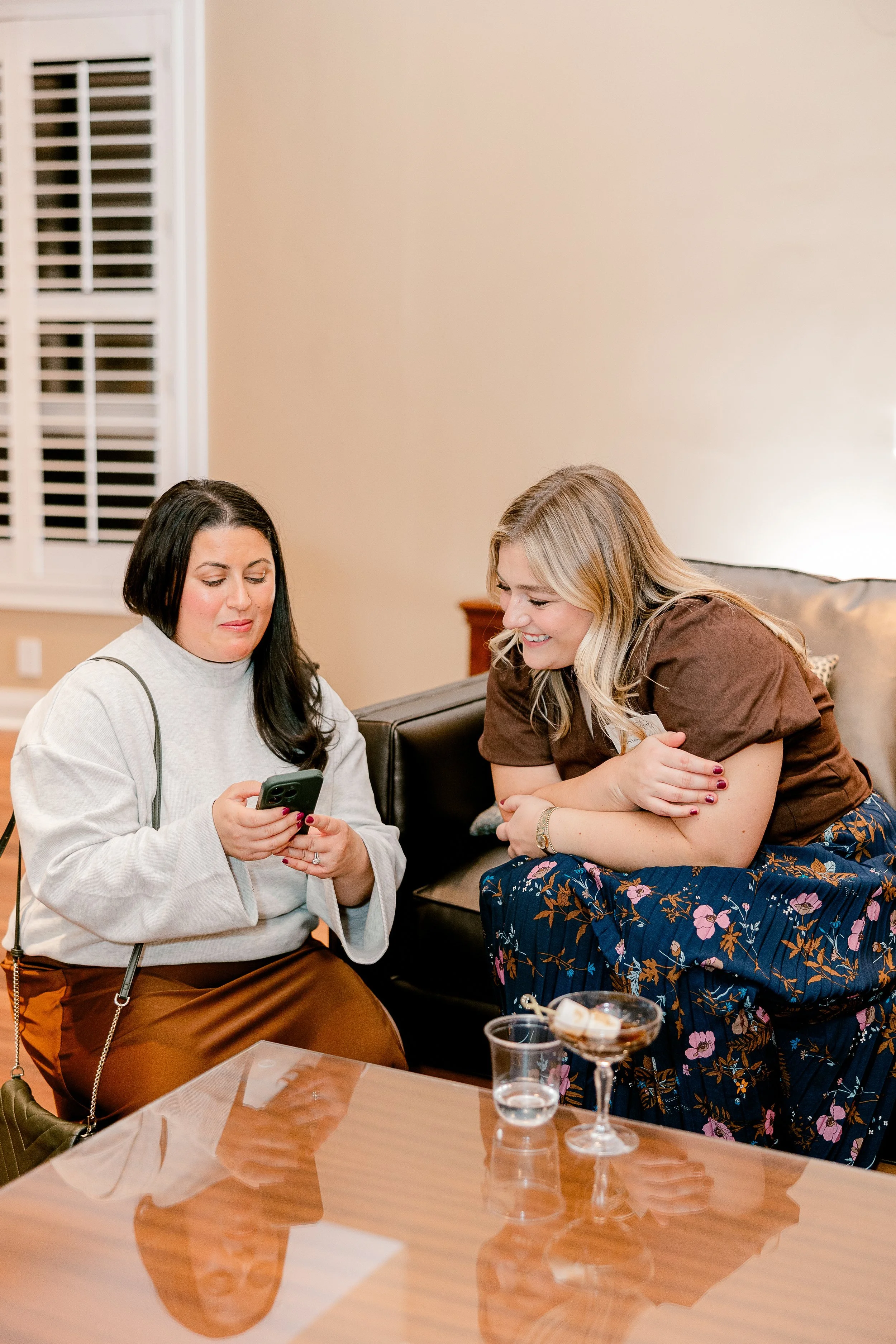 Two women sitting on a black couch in a living room, one with dark hair looking at a phone and one with blonde hair smiling, wearing a floral skirt, with drinks on a wooden table in front of them.