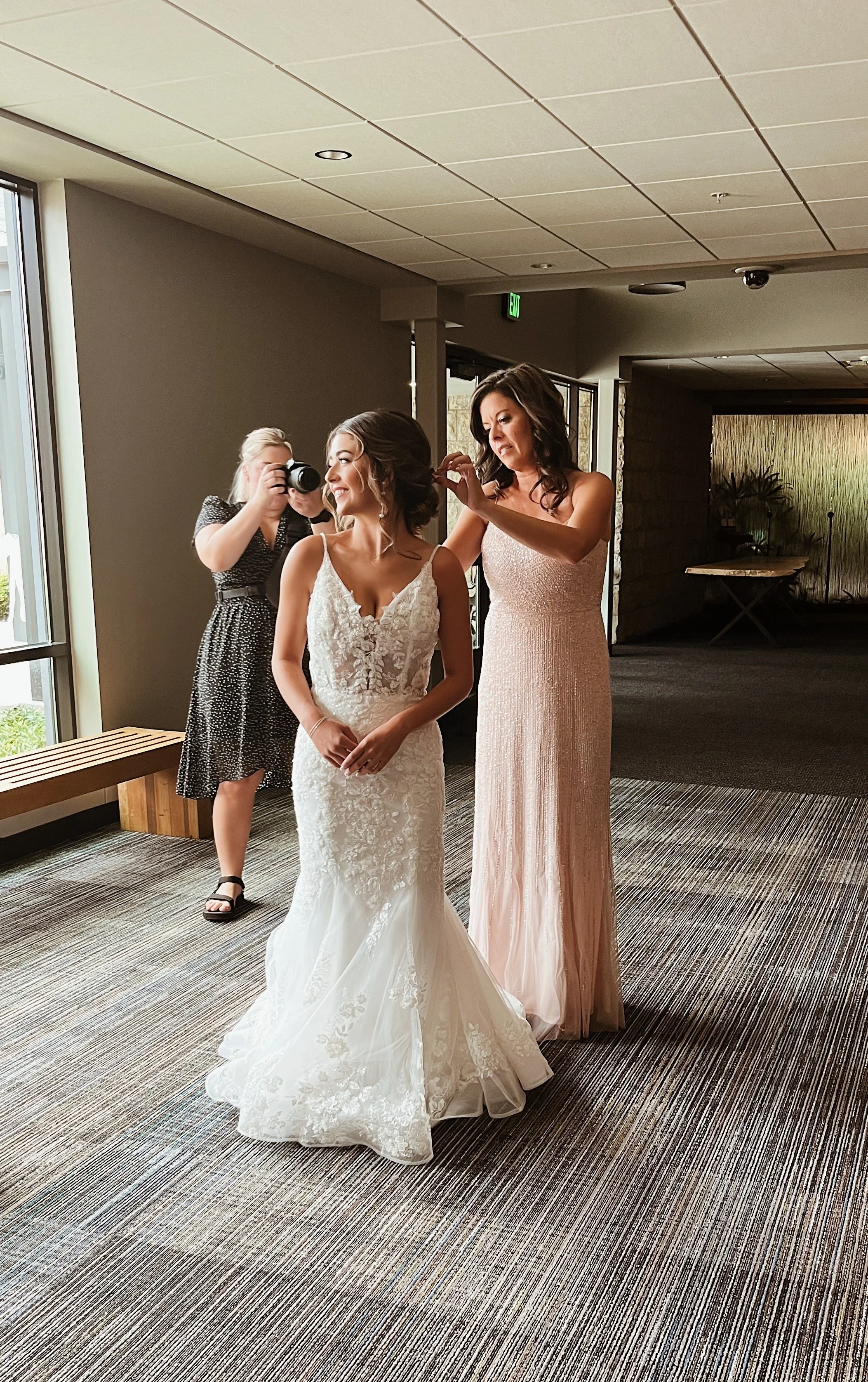 A bride in a lace wedding gown smiling as another woman in a pink dress styles her hair while a woman in black dress with a camera observes.