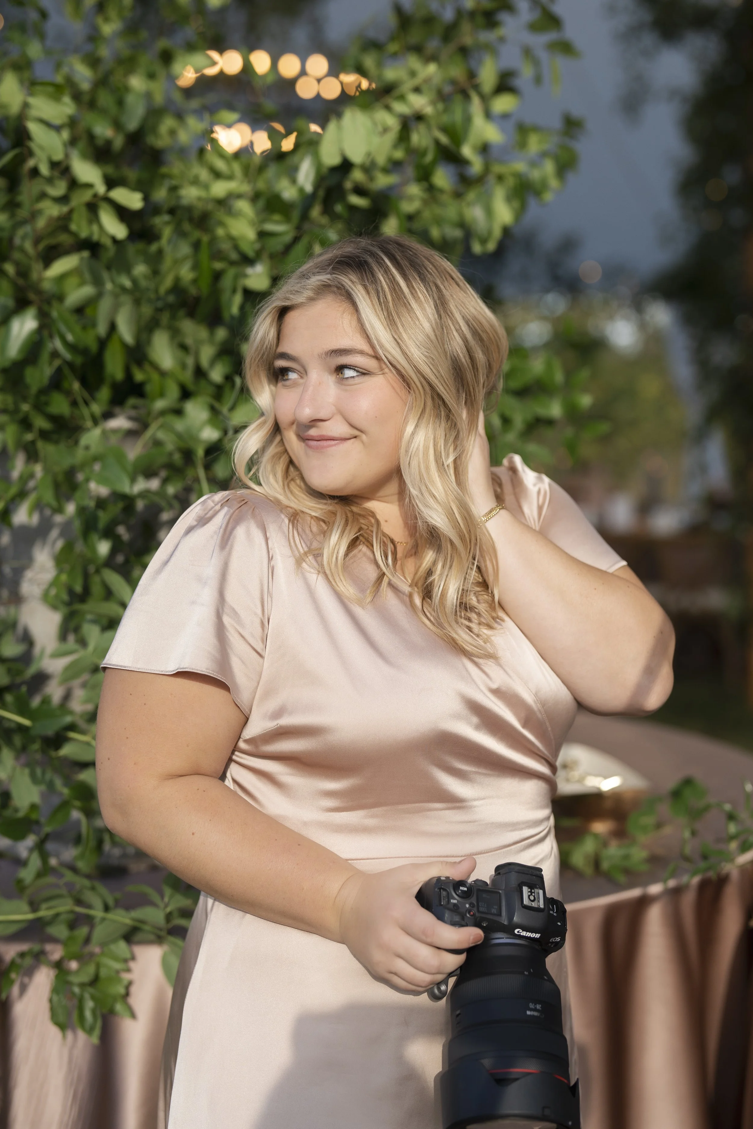 A young woman with blonde hair and a light pink satin dress holding a professional camera in an outdoor setting with green foliage and string lights in the background.