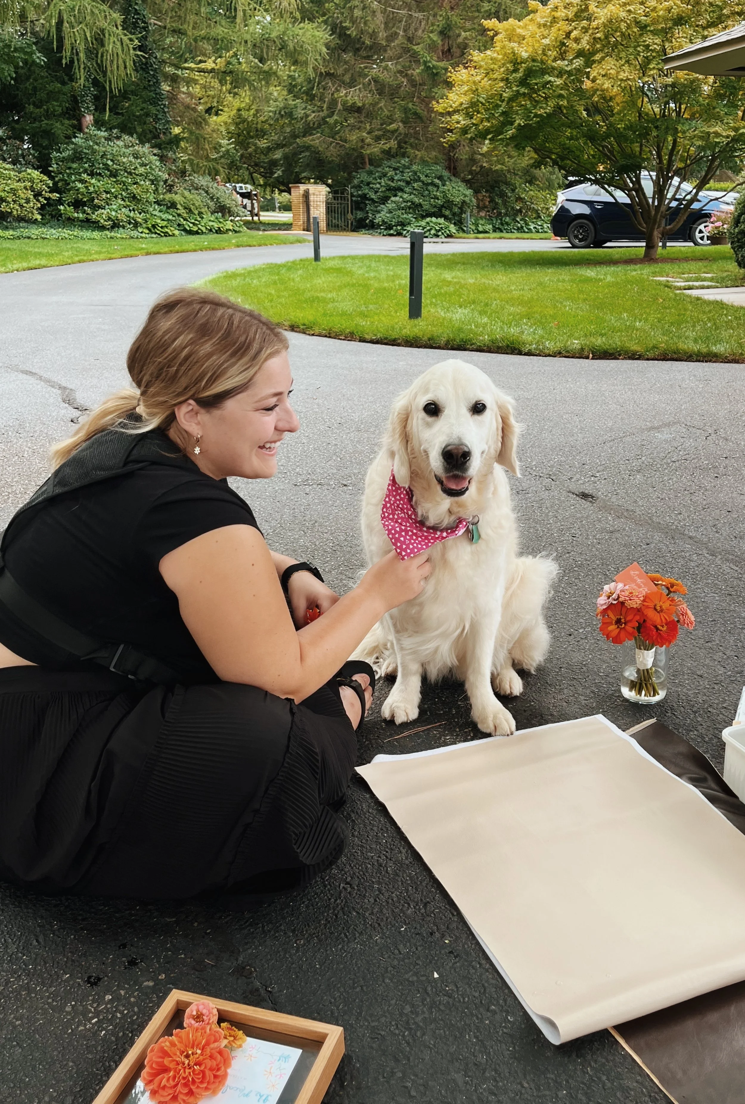 A woman with long hair and earrings is kneeling on the ground, smiling, and holding a golden retriever dog with a pink bandana around its neck. The dog is sitting on the pavement next to a small flower arrangement in a glass jar and a bouquet of oran