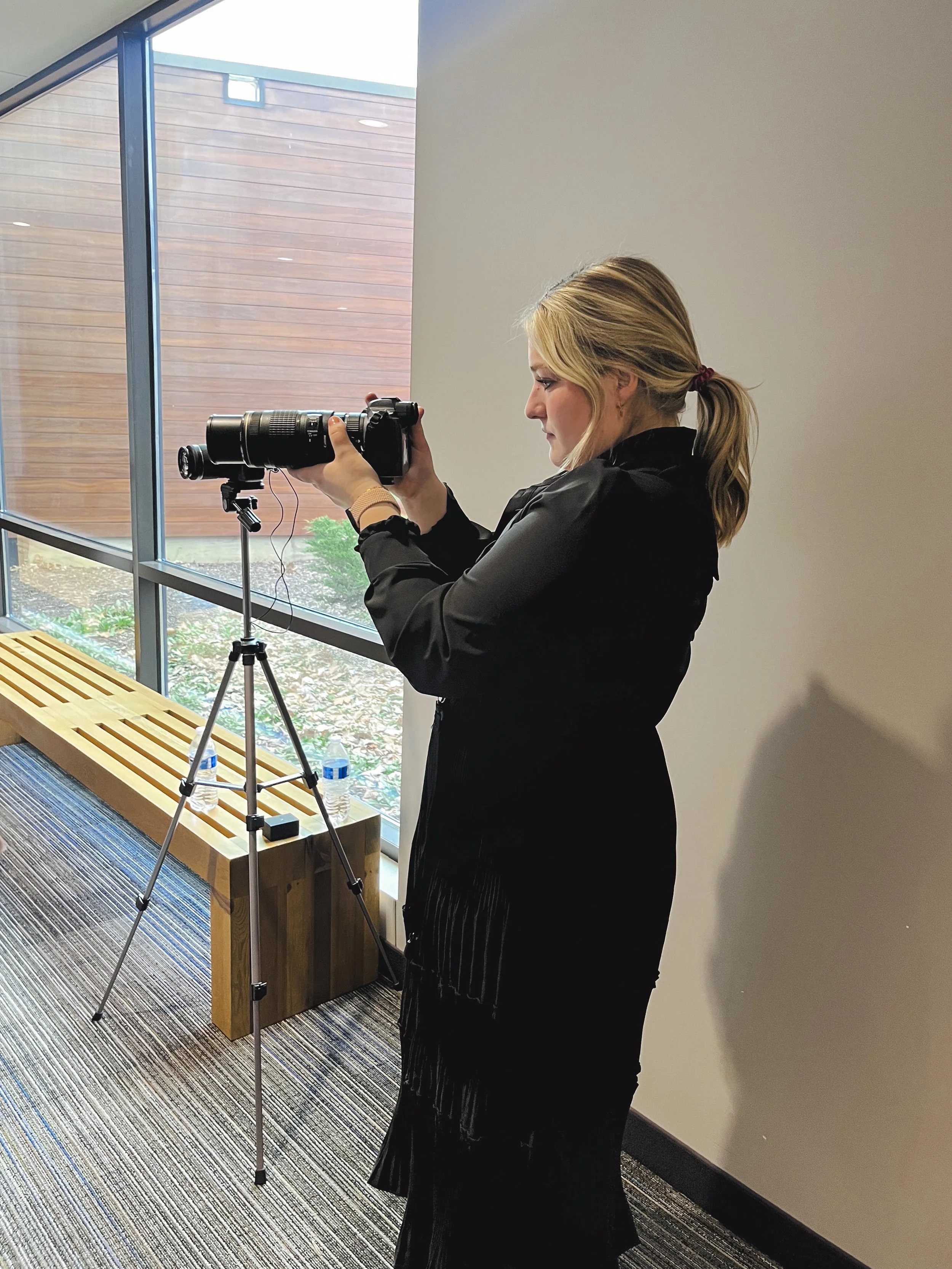 A woman with blonde hair tied in a ponytail is looking through a camera mounted on a tripod, standing near a large window with natural light coming in.