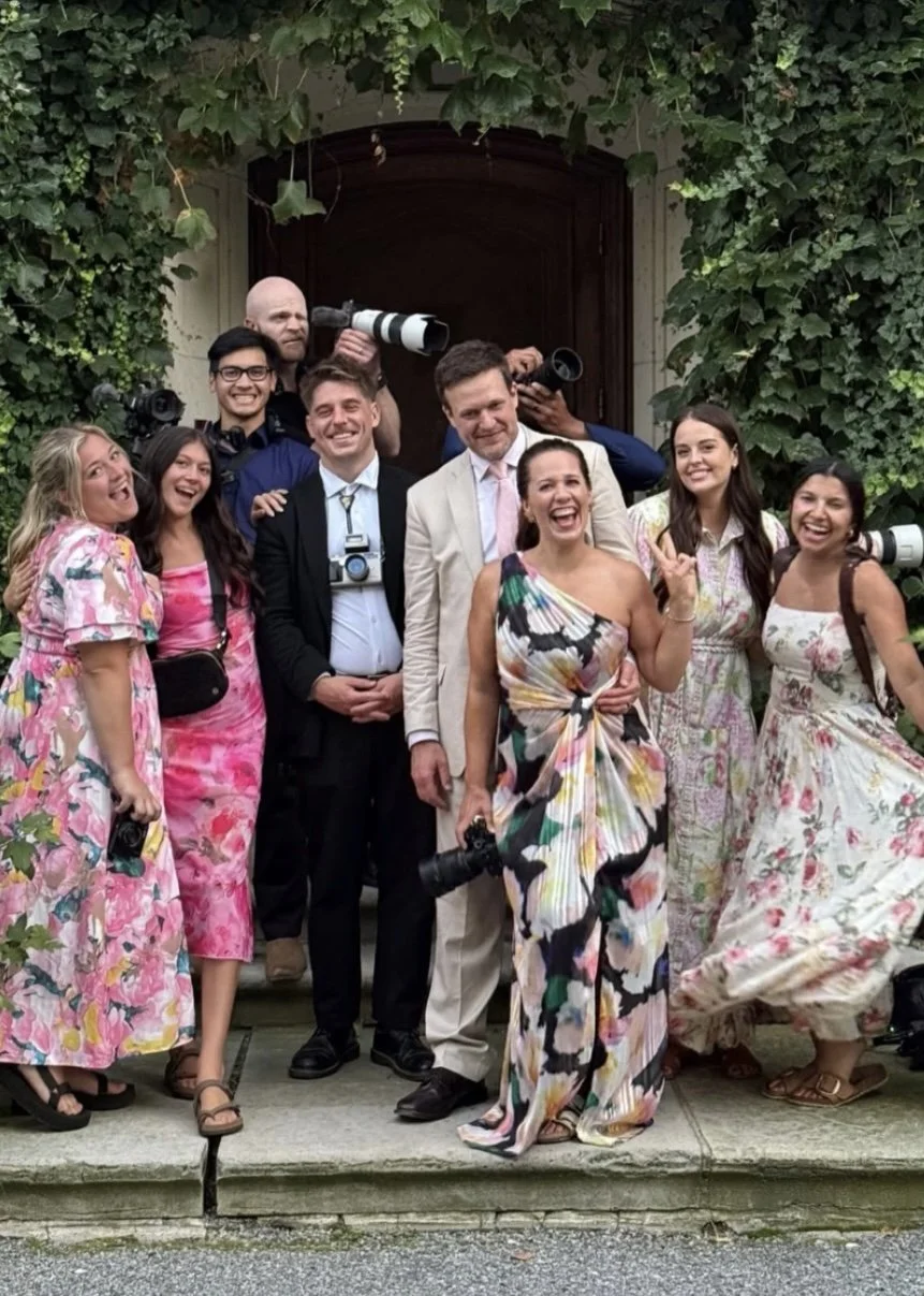 Group of people smiling and posing outside in front of an arched doorway covered with green ivy, some holding cameras and wearing colorful dresses and suits.