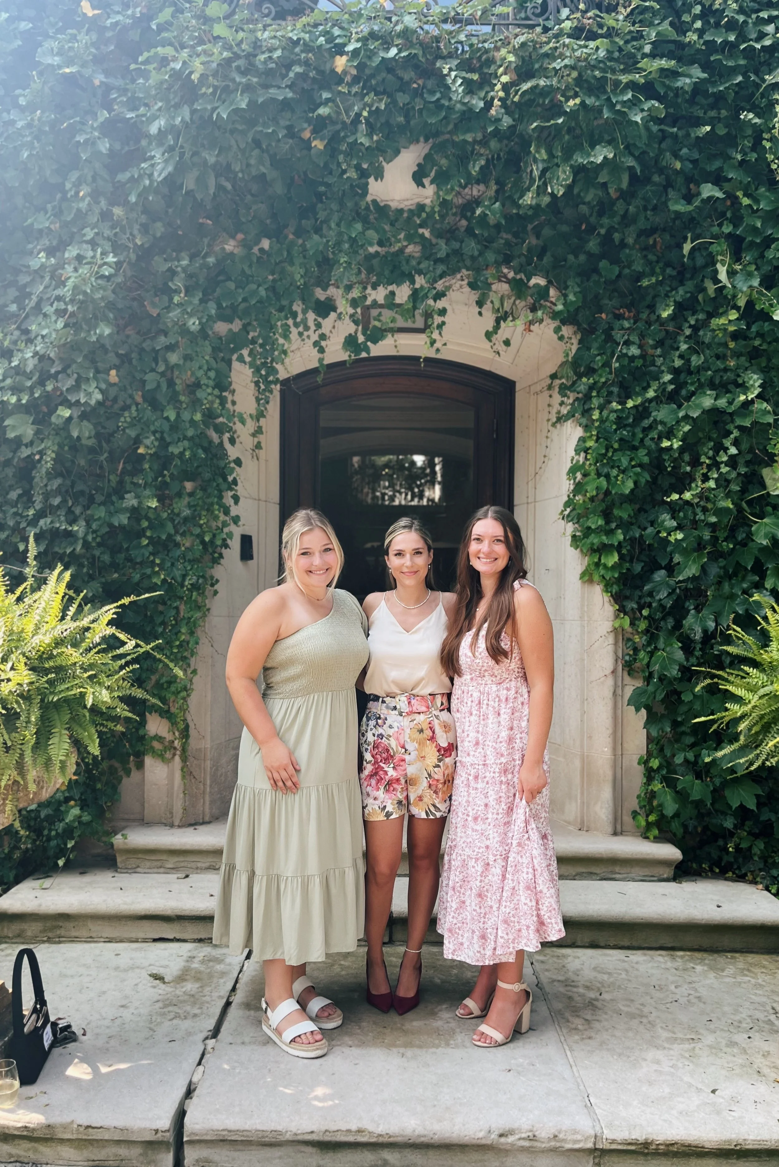 Three women standing together on a porch in front of a large archway covered with greenery, smiling at the camera.