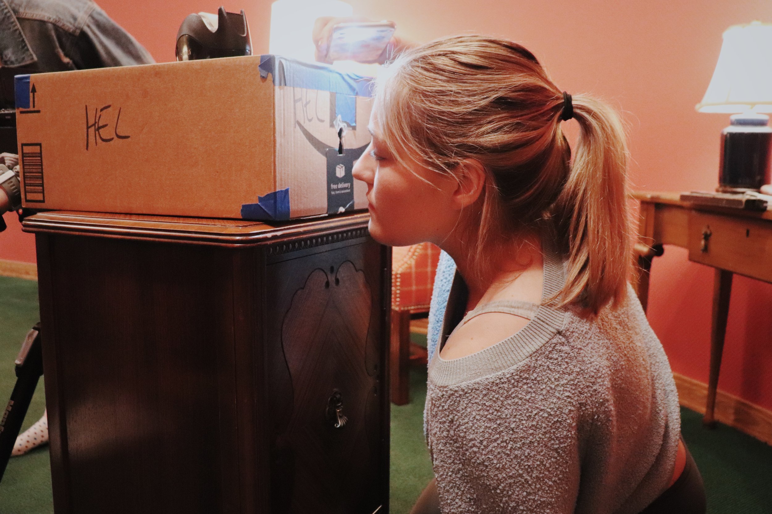 A young girl with light brown hair tied back in a ponytail, leaning her face against a cardboard box labeled 'HELL,' in a room with warm lighting, with a lamp and wooden furniture in the background.