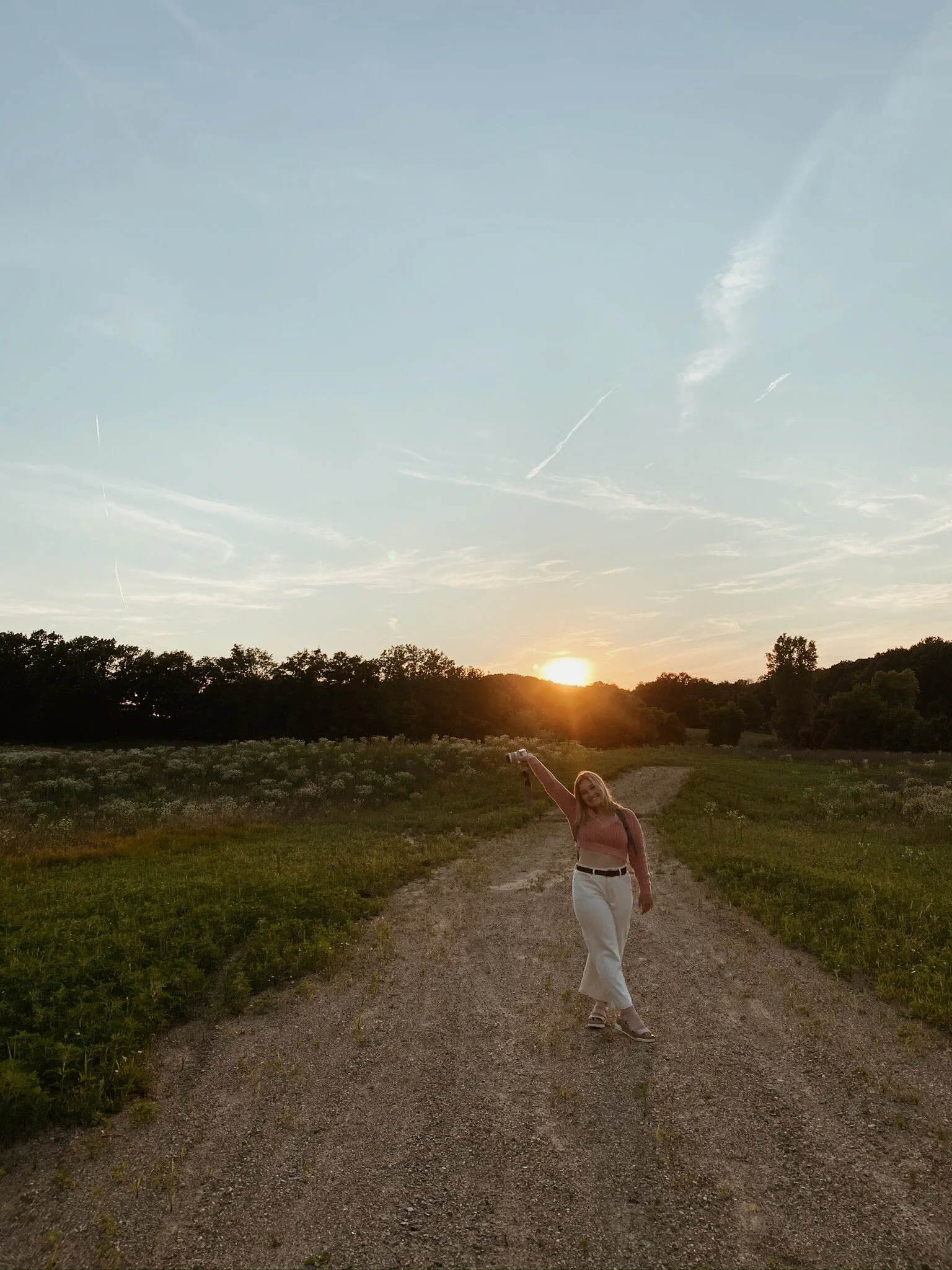 A woman walking on a dirt path during sunset in a rural area with trees on either side and a clear sky above, some clouds, and jet trails.