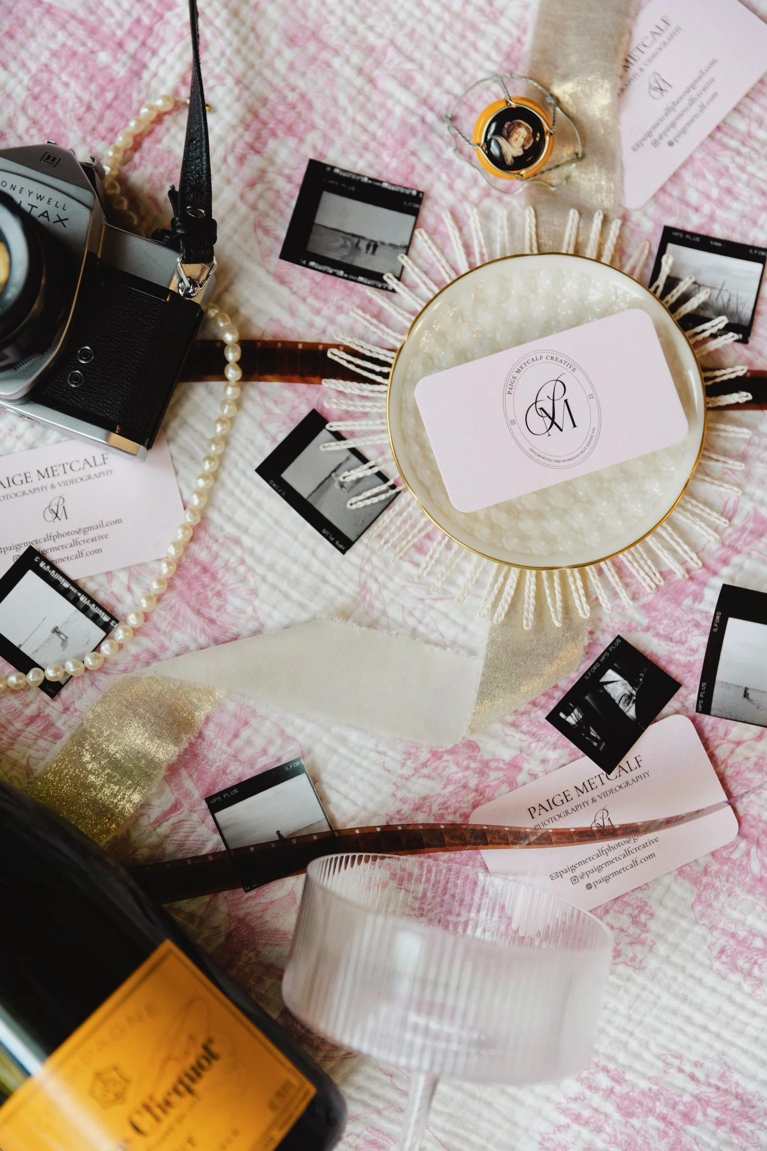 Flat lay of photography and videography equipment and accessories on a pink and white textured fabric. Includes a vintage film camera, small printed black and white photographs, business cards with name Paige Metcalf, a pearl necklace, a candle with 