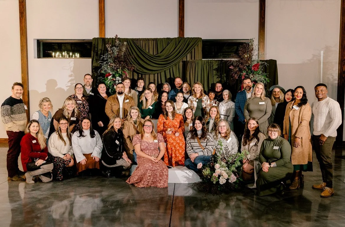 A large group of people posing together indoors in front of a decorated backdrop with flowers and green drapes.