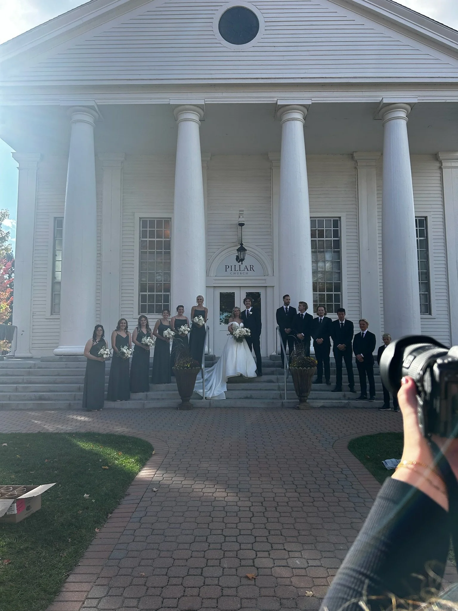 A group of people, including a bride in a white wedding dress and bridesmaids in black dresses, standing in front of a large white church with tall columns. The church's sign reads "Pillar Church." The photo is being taken by a person holding a camer