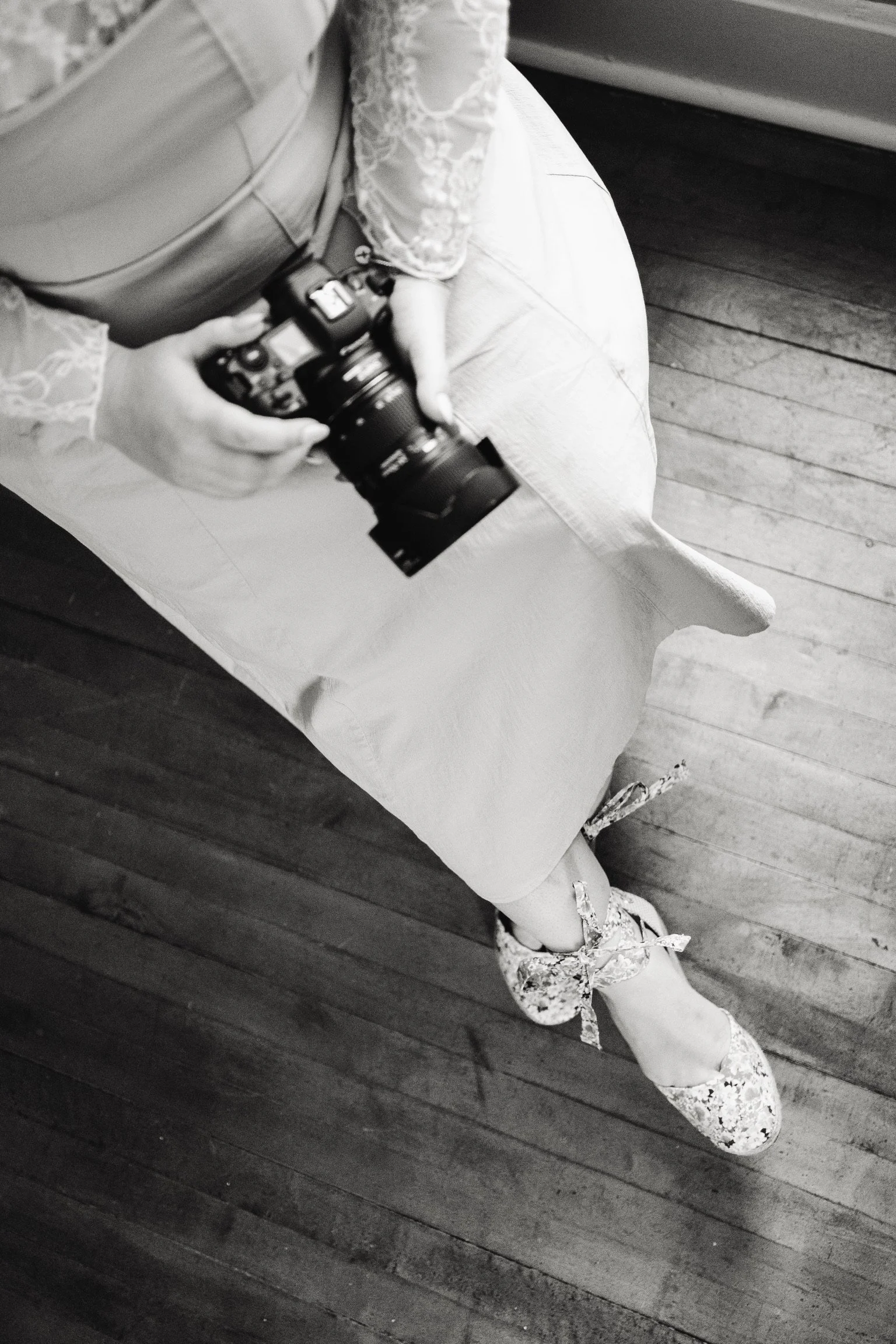 A person sitting on a bench, holding a camera. The person is wearing lace shoes, a lace skirt, and a top with lace sleeves. The photo is in black and white, taken from an overhead angle, showing the person's legs, feet, and part of the camera. The fl