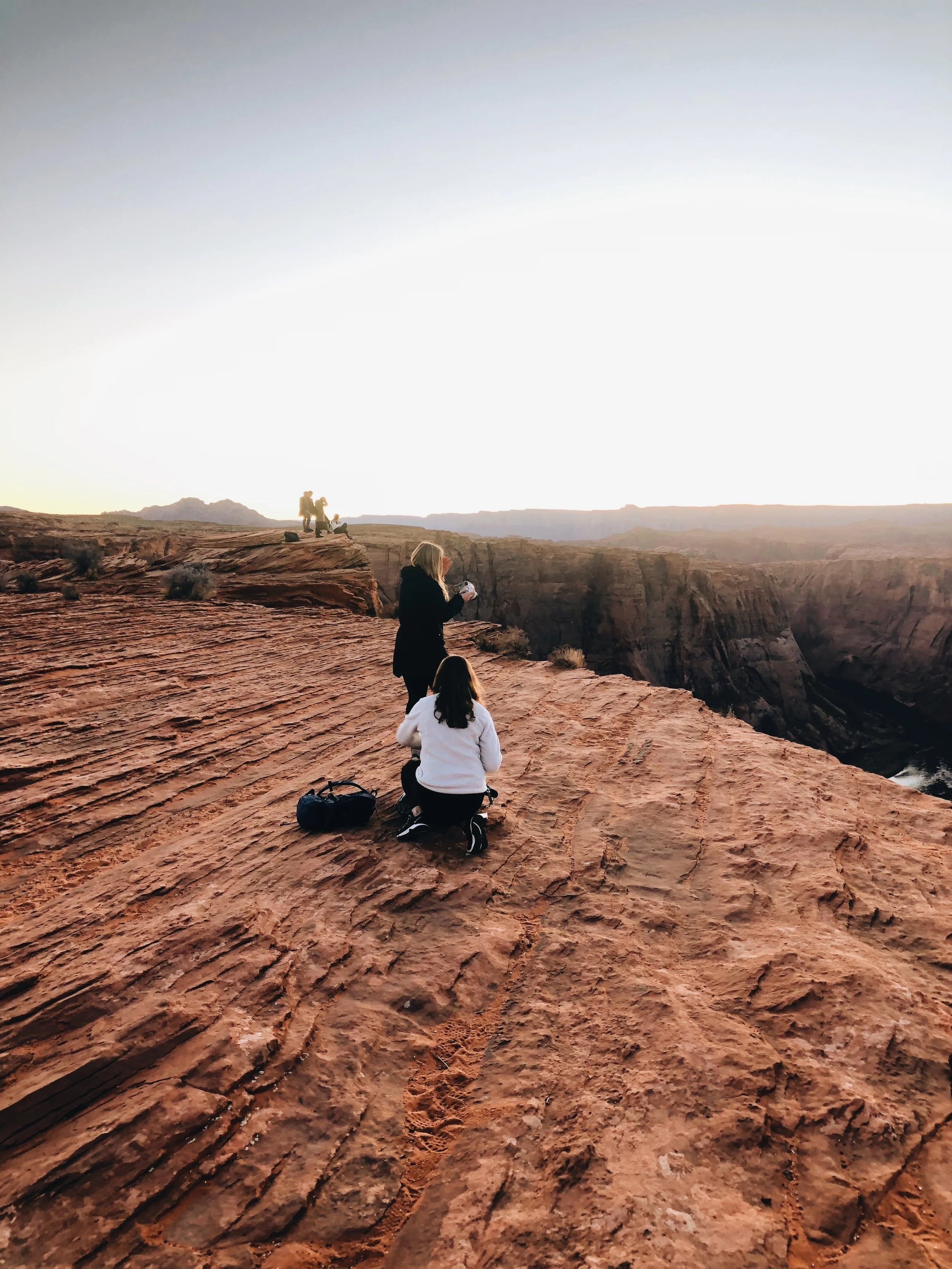 People sitting and standing on red rock cliffs at sunset in a canyon, with a woman holding a camera in the foreground.
