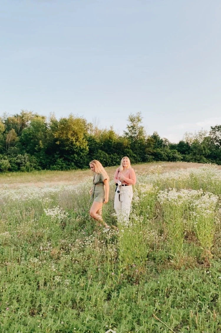 Two women walking in a grassy field with white flowers, with trees and a clear blue sky in the background.