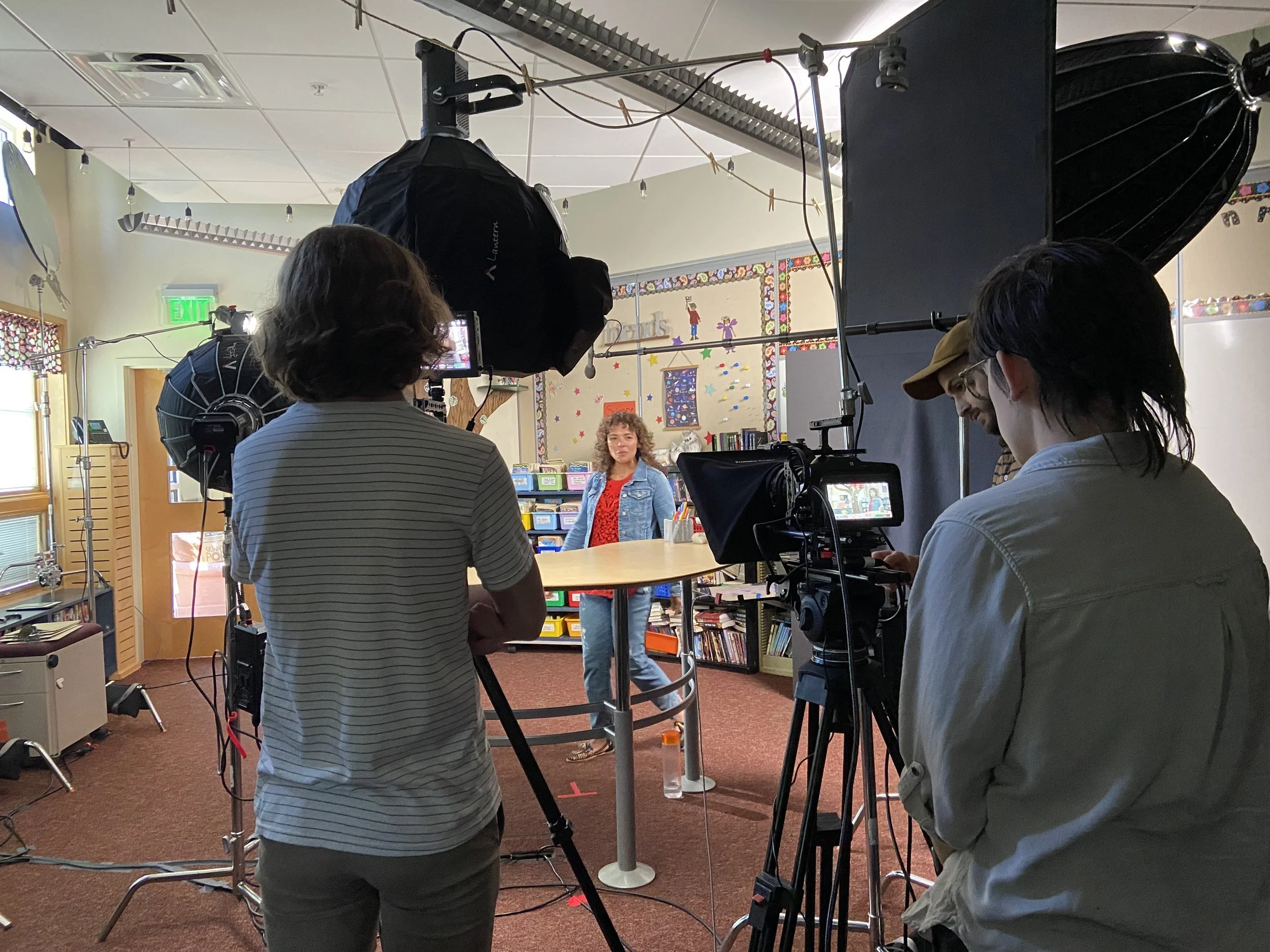Behind the scenes of a video shoot in a room with bookshelves and children's artwork. Production crew operating cameras and lighting with a woman standing in front of a colorful background.