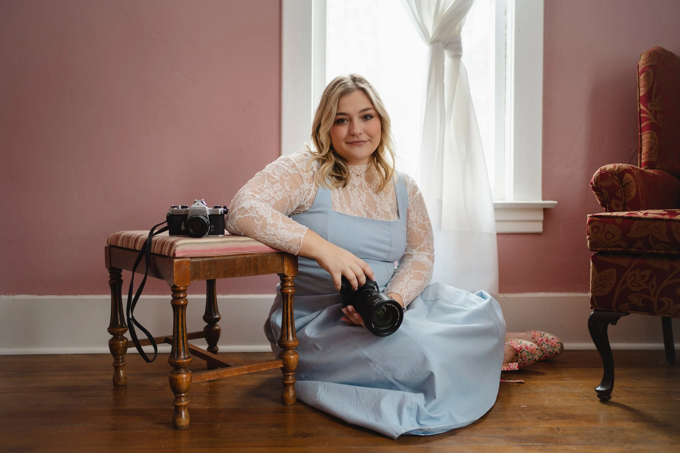 A young woman with blonde hair wearing a lace top and a light blue dress, sitting on the wooden floor next to a vintage camera on a table, holding a larger camera, in a room with pink walls, wooden floors, and a window with draped curtains.