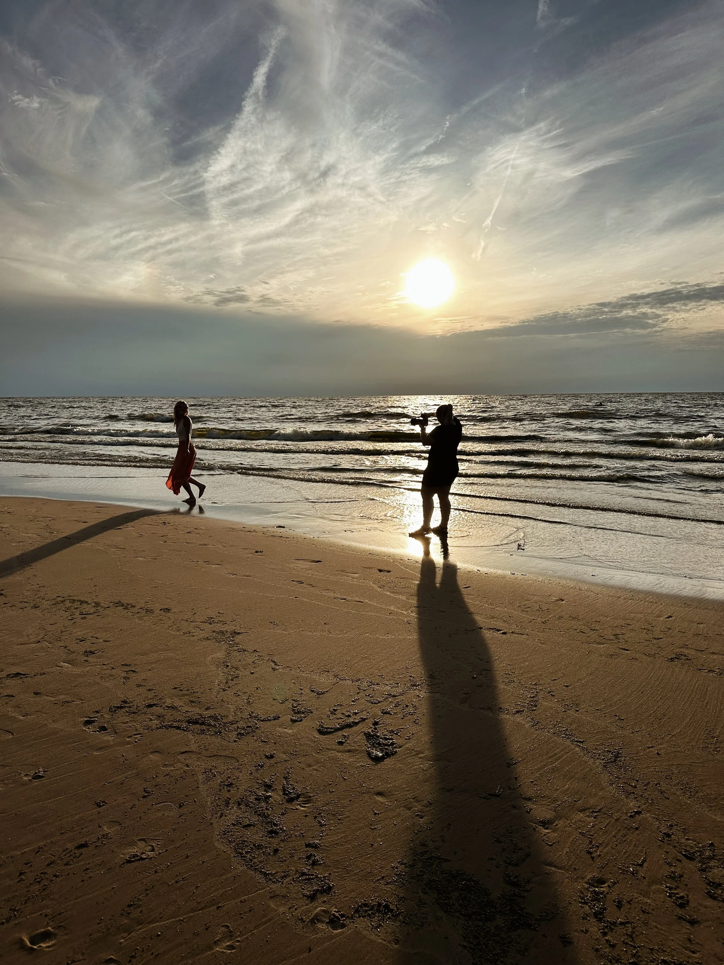 The image depicts a sunset at the beach with two people taking photographs of the scene. The sky is partially cloudy with the sun low on the horizon. The beach has footprints and a long shadow cast by one of the people.