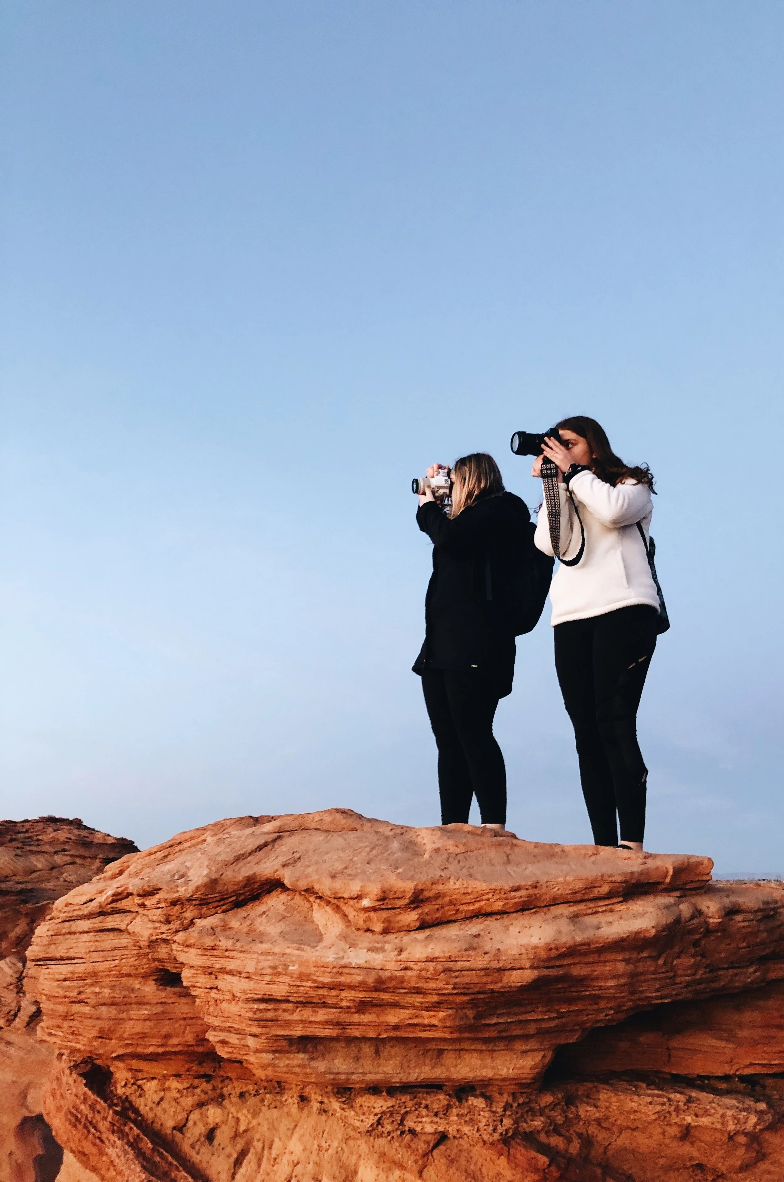 Two women standing on a rock formation, each holding a camera and taking photographs. The sky is clear and blue.