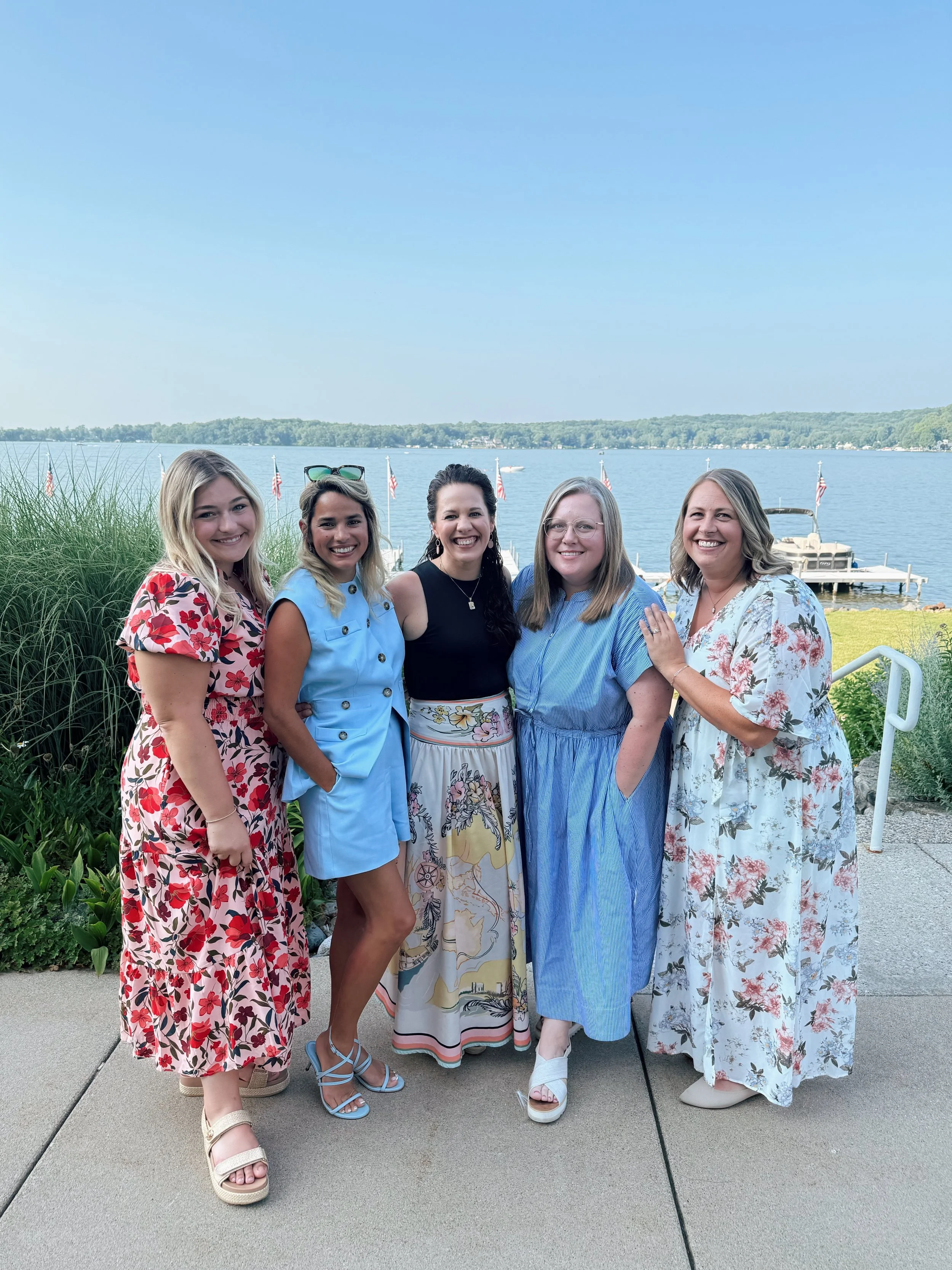 A group of five women standing outdoors near a body of water with sailboats and American flags in the background. They are smiling and dressed in colorful, floral, and striped dresses.