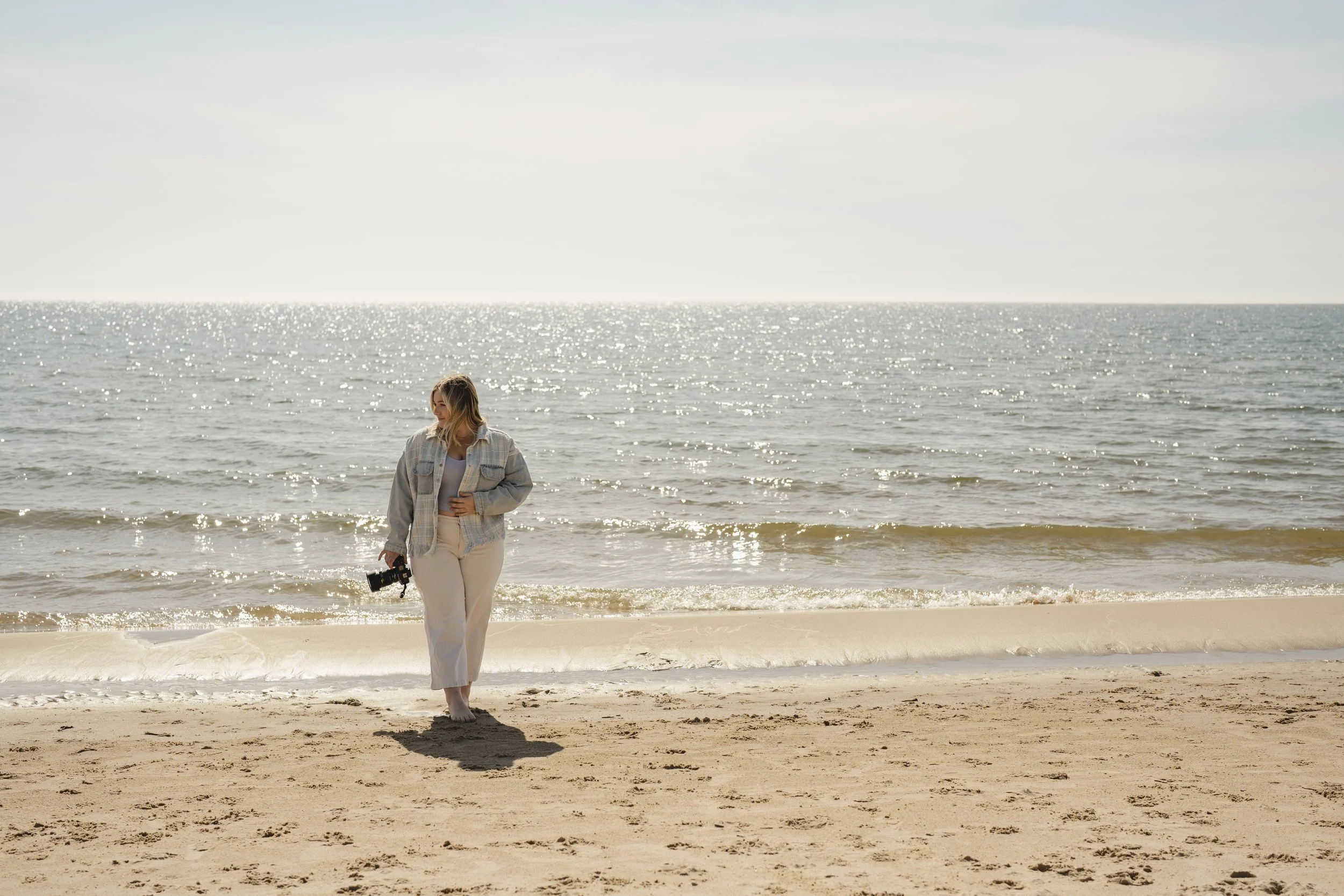 A woman walking barefoot on the beach, holding a camera, with the ocean and sky in the background.