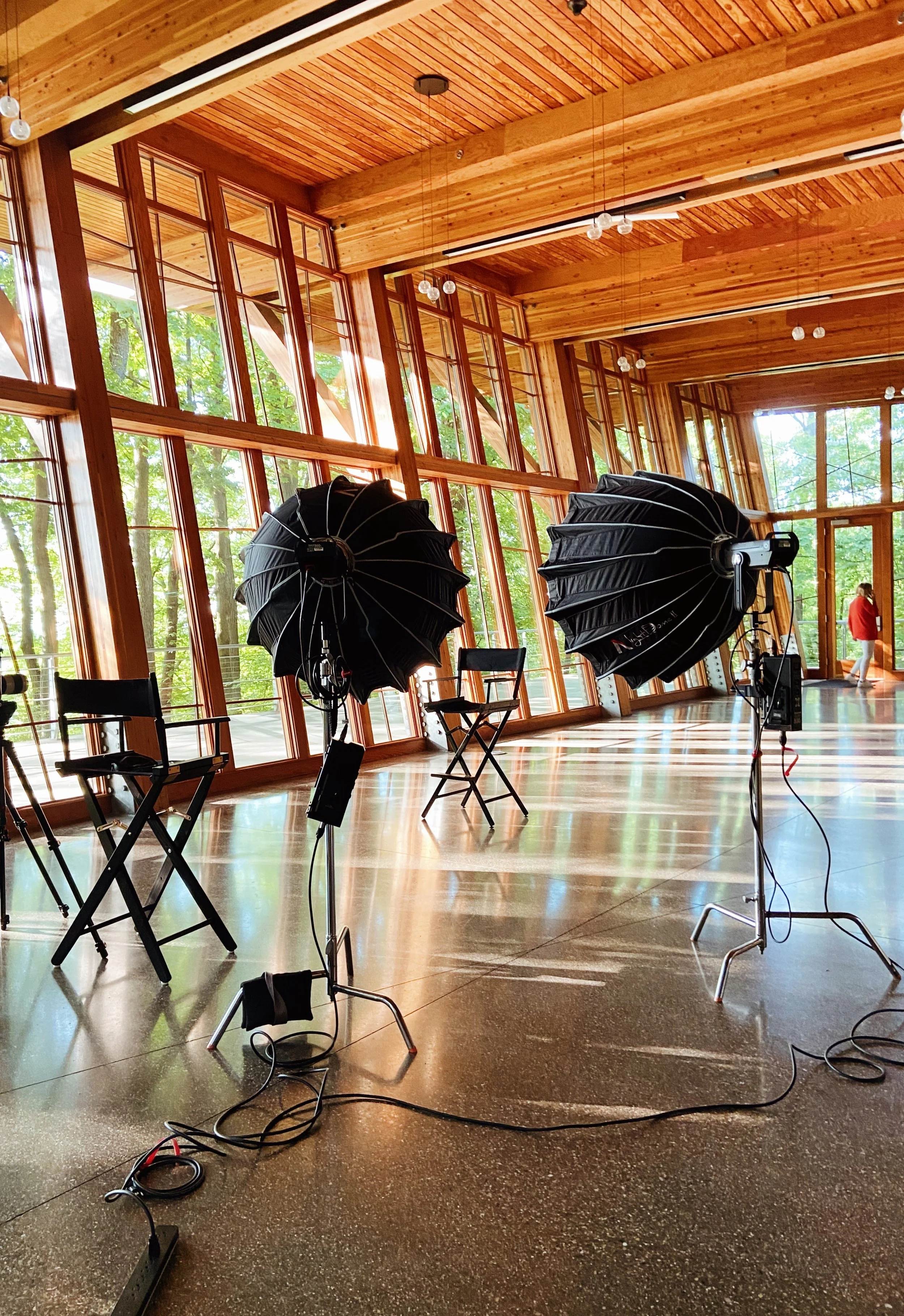 Inside a wooden studio with large windows, photography equipment including softbox lights, chairs, and power strips are set up. A person in a red jacket is visible in the background near the windows.