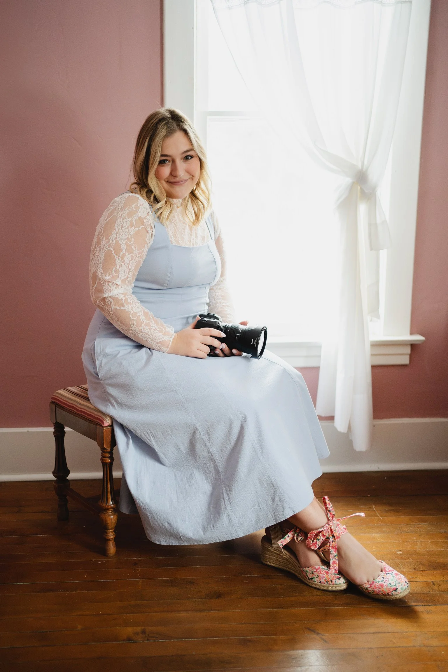 A young woman sitting on a wooden stool in a room with pink walls and wooden flooring, holding a camera, smiling, dressed in a light blue dress with lace sleeves, wearing floral wedge slippers, near a window with white curtains.