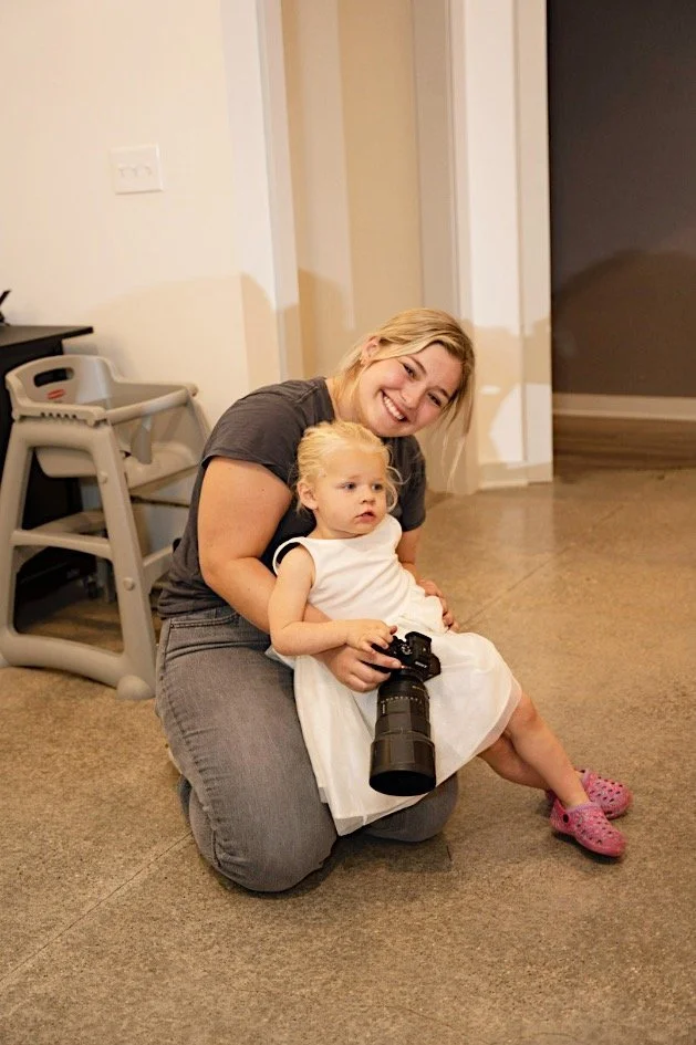 A woman smiling and hugging a young girl sitting on her lap, holding a camera with a large telephoto lens, in a room with beige carpet and white walls.