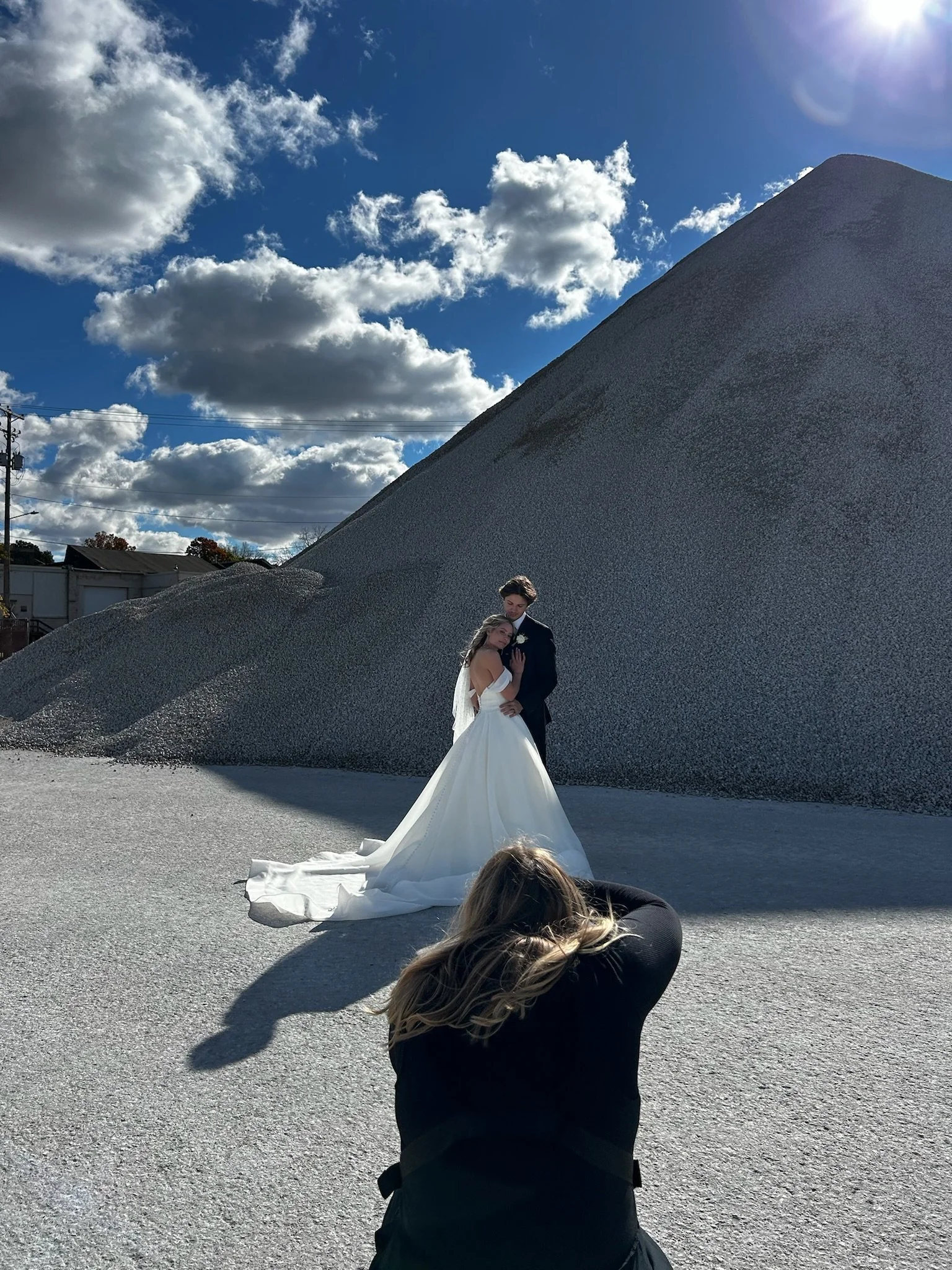 A bride and groom pose for a wedding photo outdoors in front of a large gravel mound under a blue sky with scattered clouds. A woman in black kneels in the foreground, capturing the moment, while a photographer with blonde hair lies on the ground tak