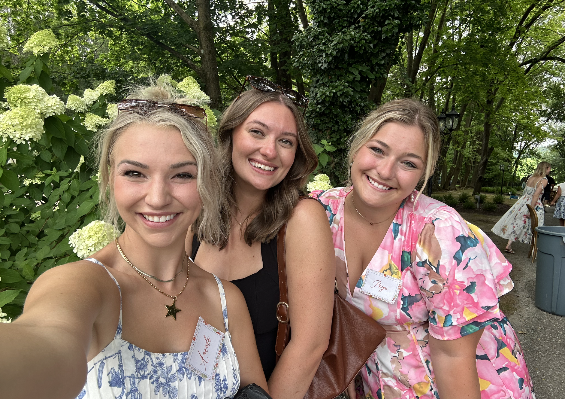 Three women smiling and taking a selfie outdoors, surrounded by green trees and bushes, with other people in the background at a gathering or event.