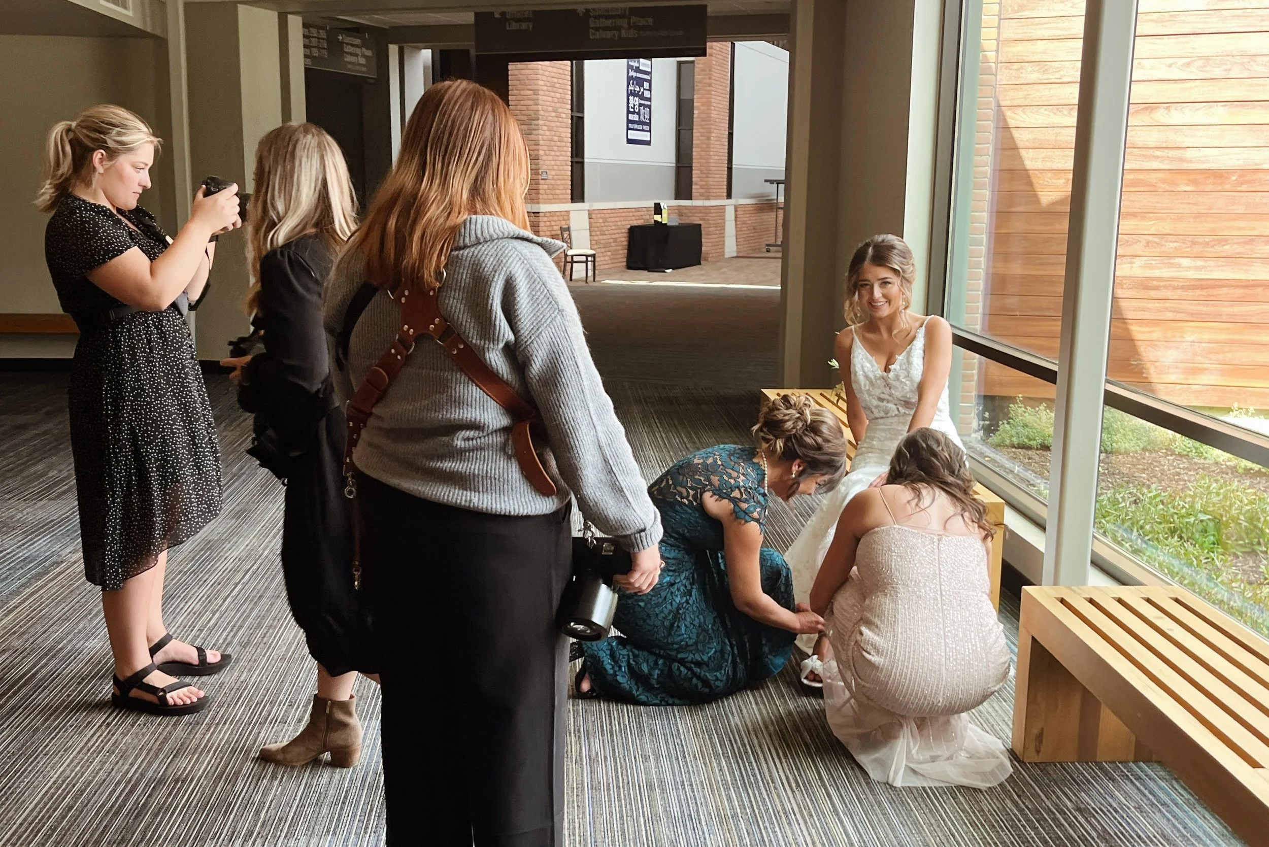 Group of women, some standing and some kneeling, gathered by a large window while a bride in a white wedding dress is seated near the window smiling. The scene appears to be a wedding-related activity, possibly a pre-ceremony moment or photo shoot.