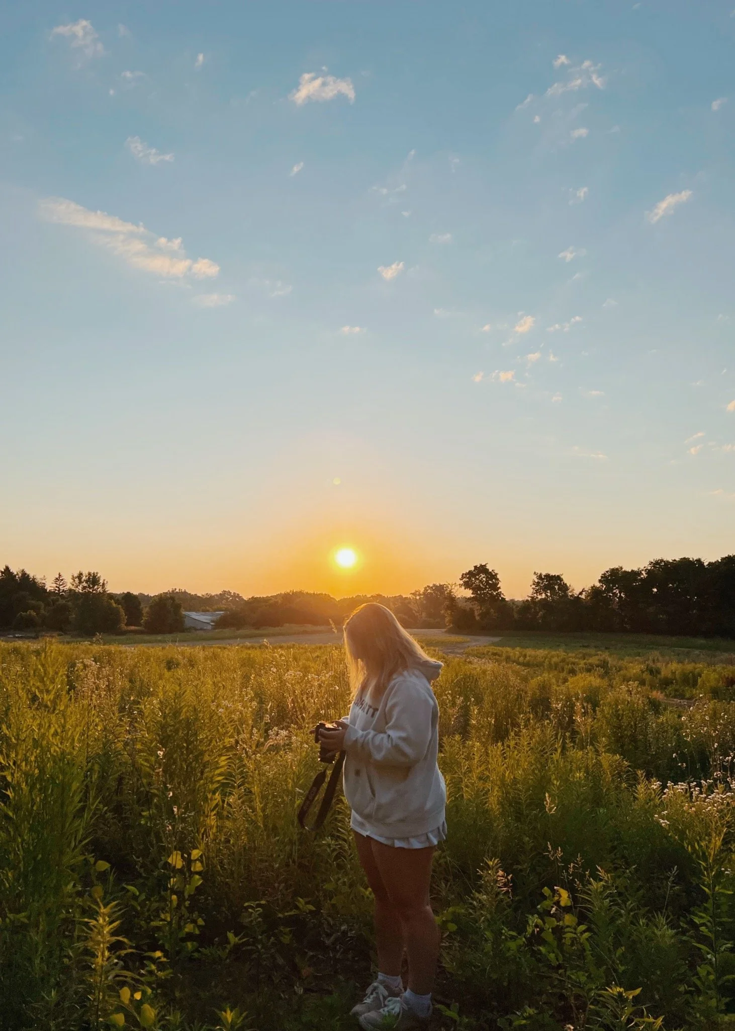 A person standing in a field of tall grass at sunset, holding a camera or phone, with trees and a clear sky with some clouds in the background.