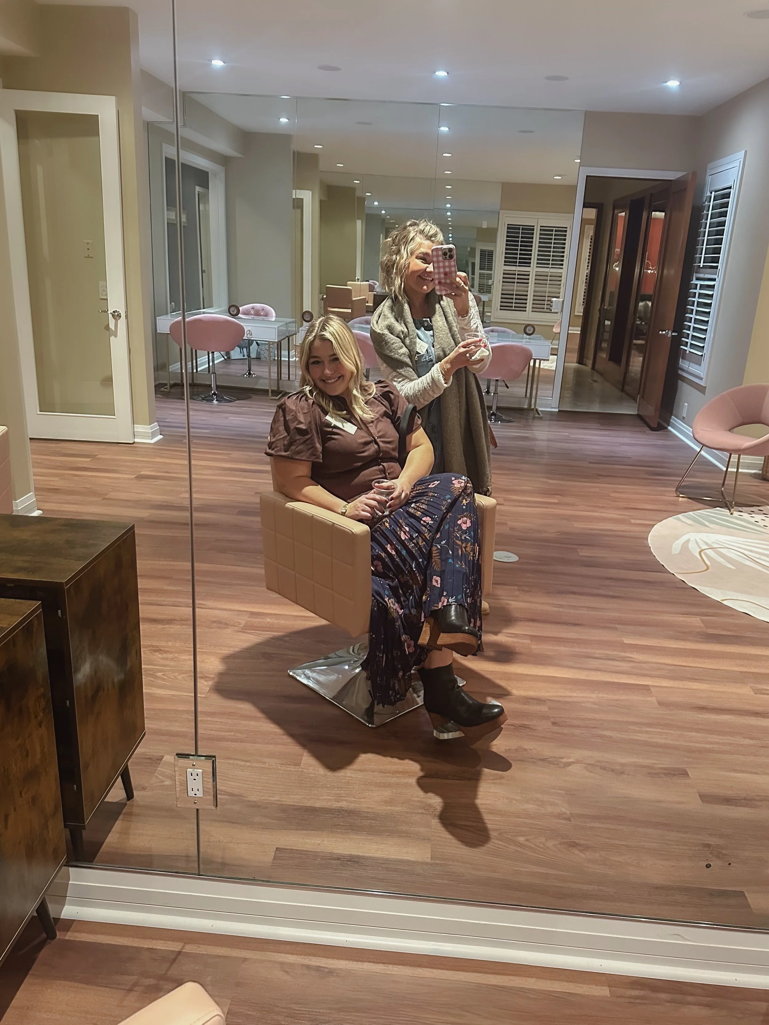 Two smiling women taking a selfie in front of a large mirror in a stylish room with wood flooring and pink chairs.