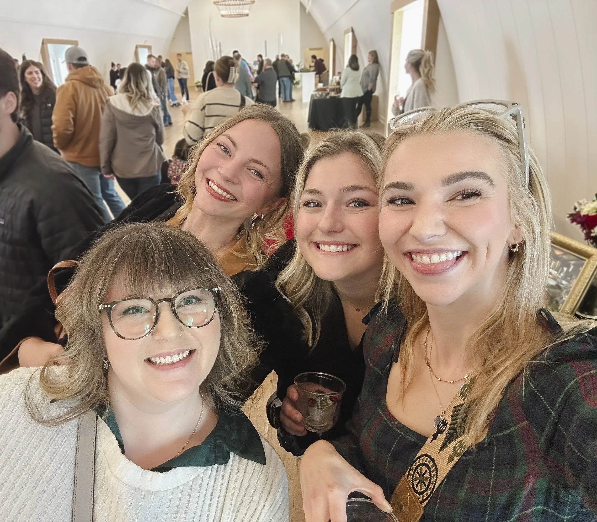A group of women and men at an indoor social event, smiling for a selfie. The background shows more people mingling and a reception area with tables and decorations.