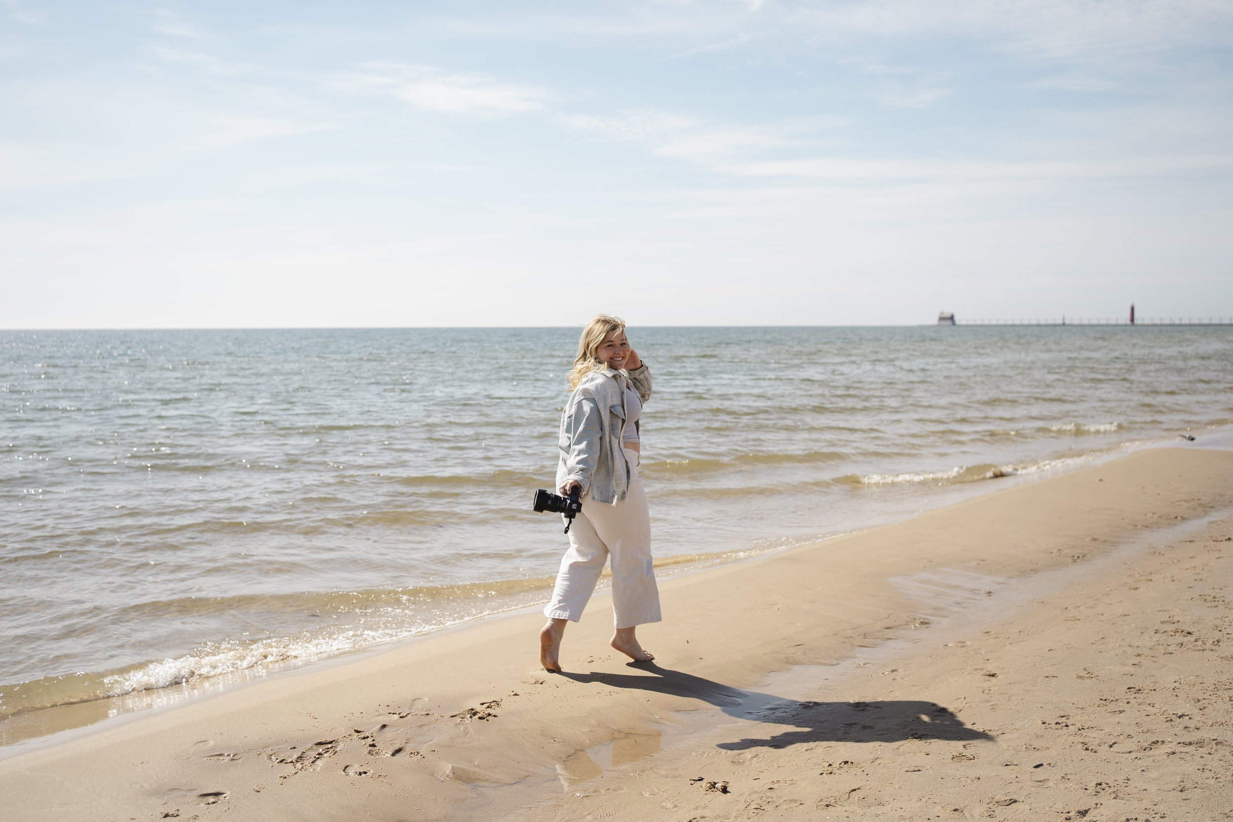 A woman with blonde hair walking at the beach, holding a camera, with ocean waves and lighthouse in the background.