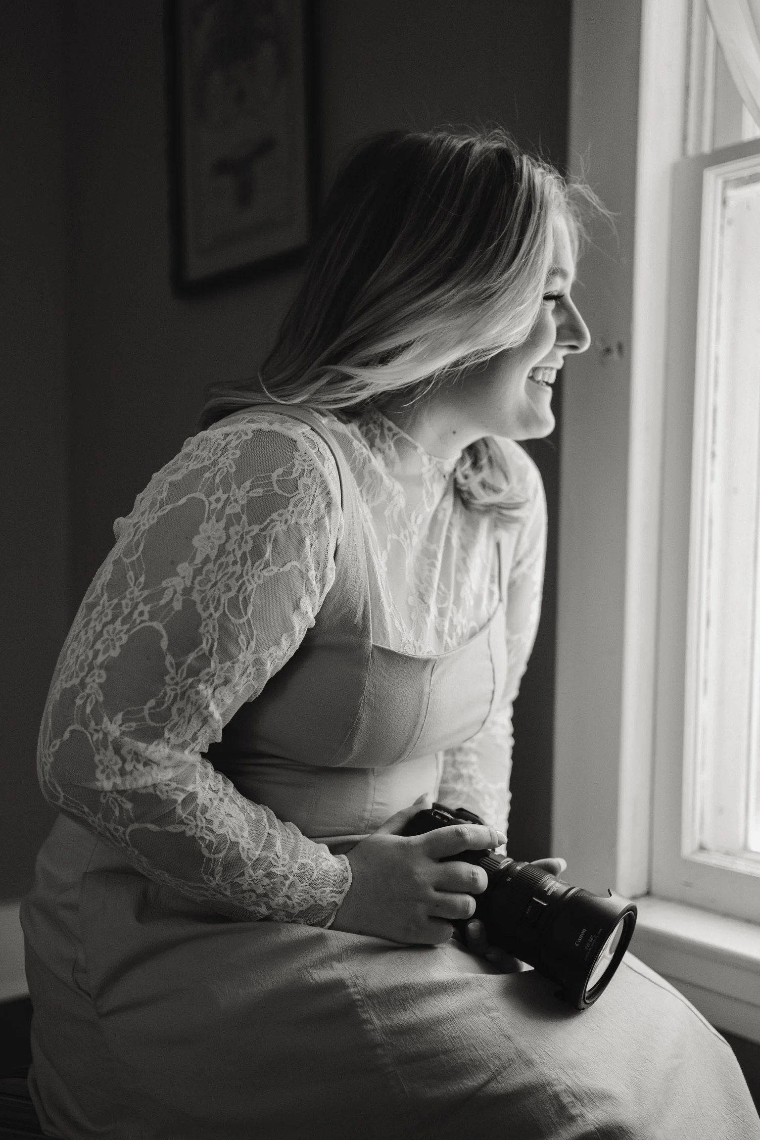 A woman in a lace top sitting near a window holding a camera, smiling and looking outside.