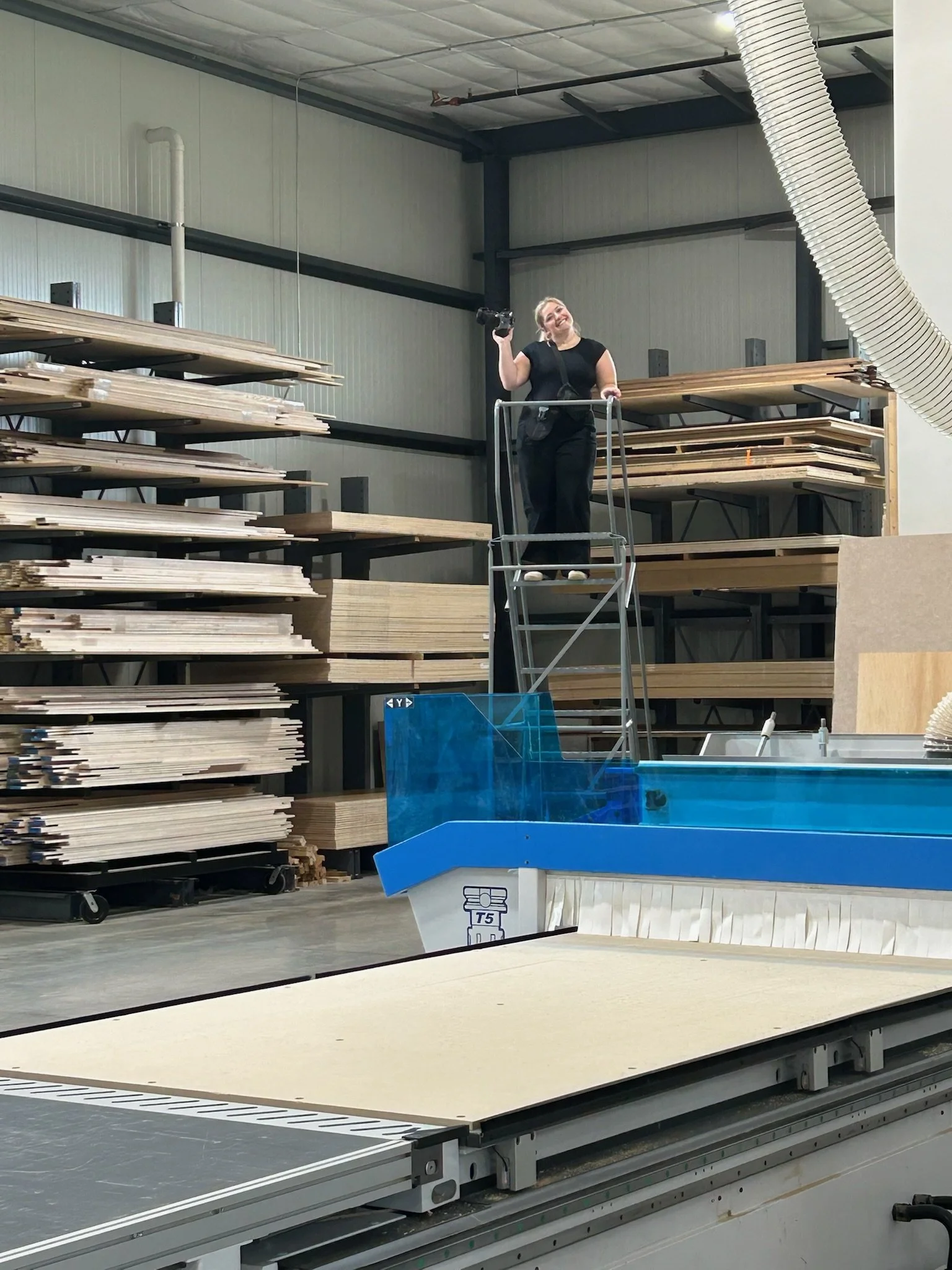 A woman standing on a platform in a warehouse filled with stacks of wooden boards, holding a camera and smiling at the camera. There is a blue and white object in the foreground, with a large flexible duct hanging from the ceiling.