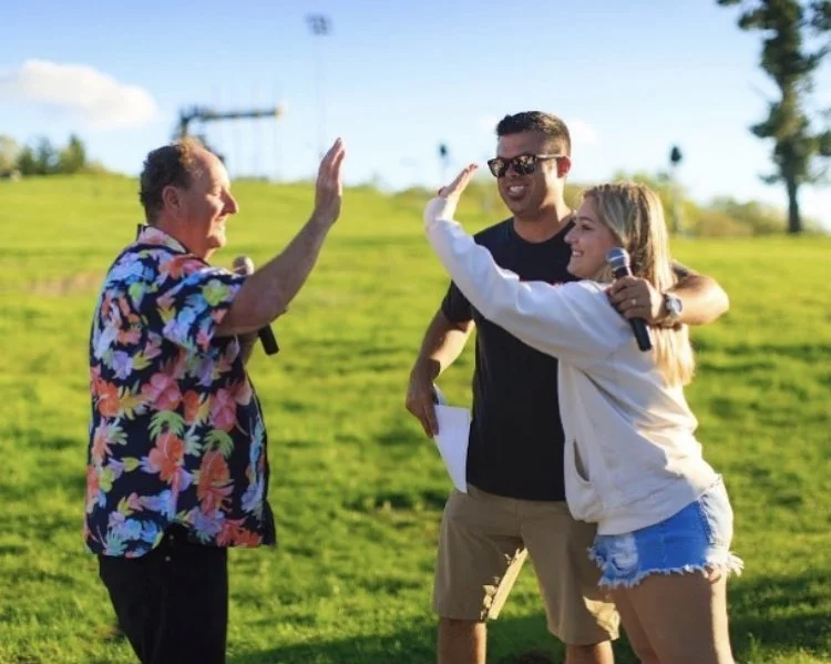Two women and one man outdoors on a grassy field, high-fiving each other, with the women holding microphones, indicating a celebration or outdoor event.