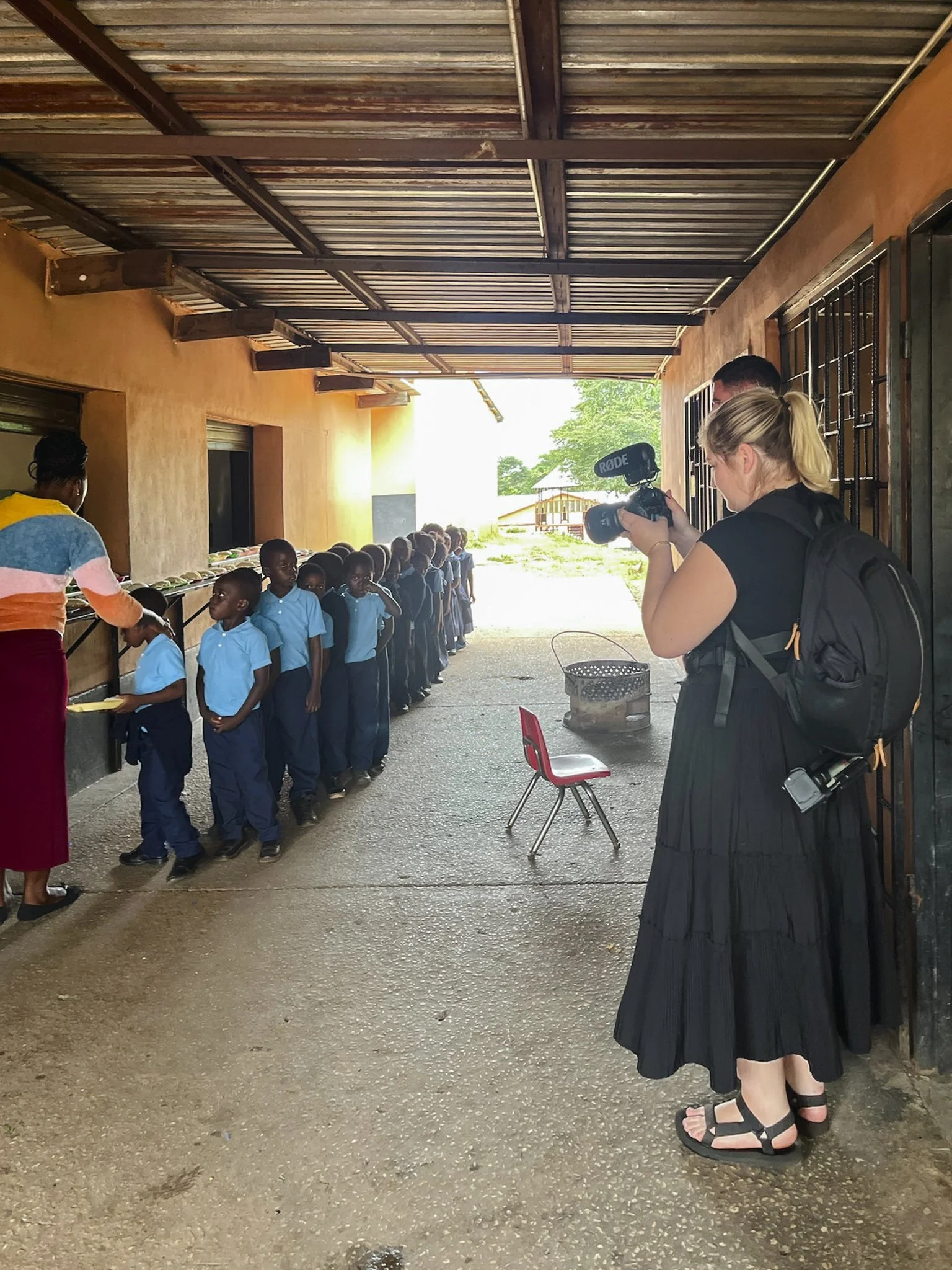 A group of children in blue uniforms standing in a line under a covered outdoor area, with a woman filming them with a camera. The woman has blond hair in a ponytail, and is wearing a black dress, sandals, and a backpack.