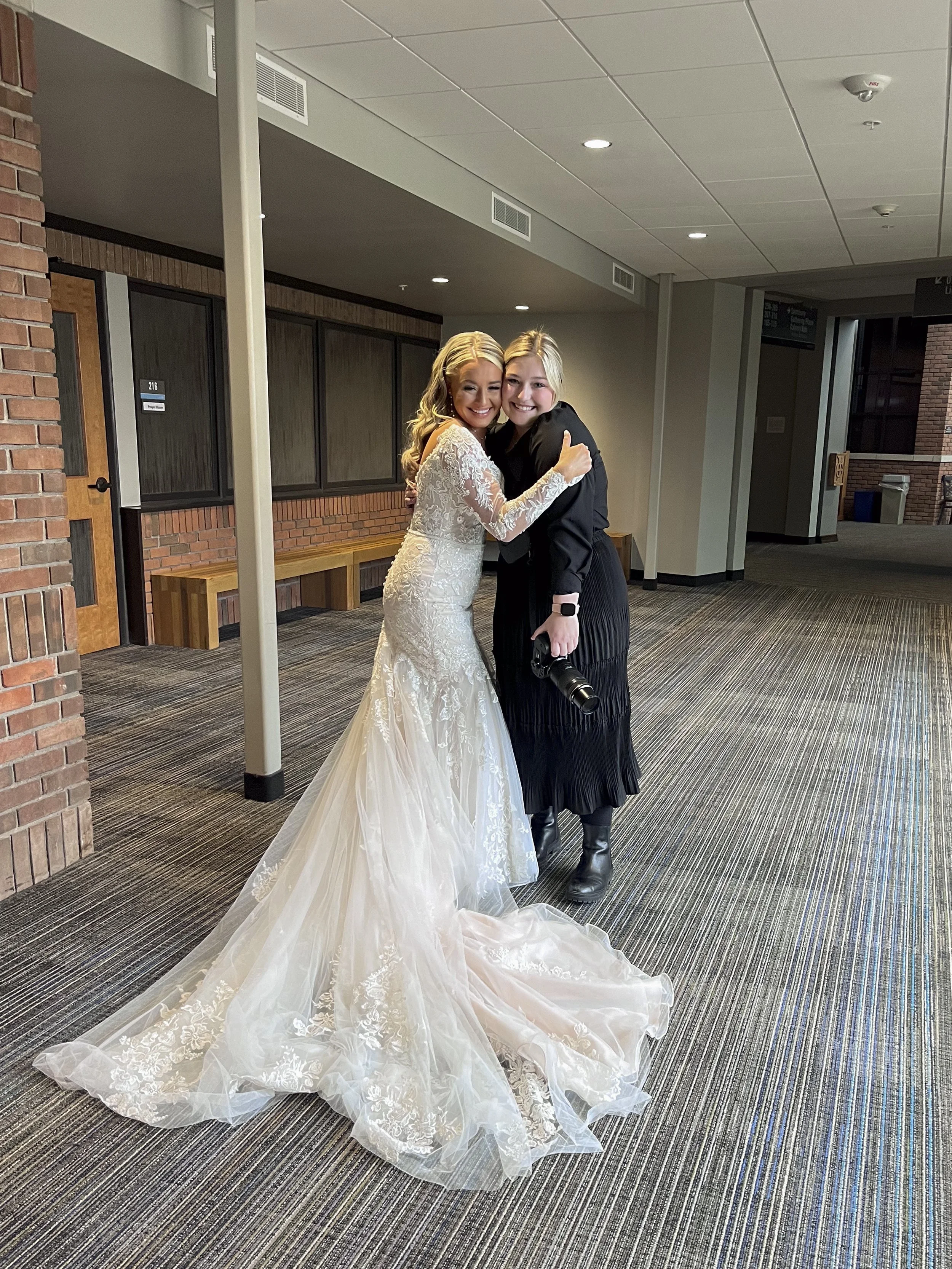 A bride and a woman are hugging and smiling in a hallway, with the bride in a white wedding dress with lace details and a long train, and the woman in a black outfit holding a camera.