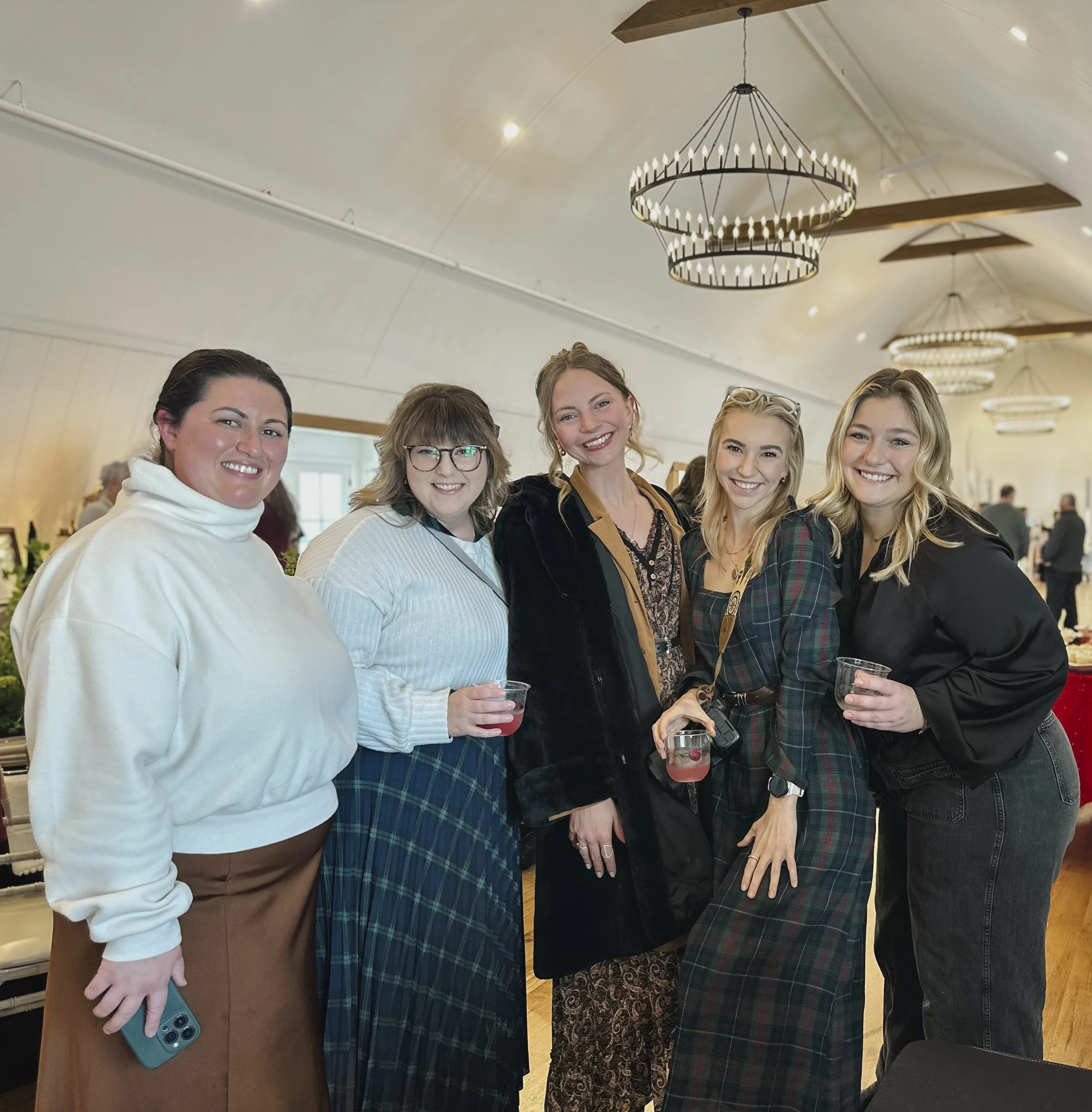 Group of women smiling at a gathering, some holding drinks, in an indoor setting with chandeliers hanging from the ceiling.