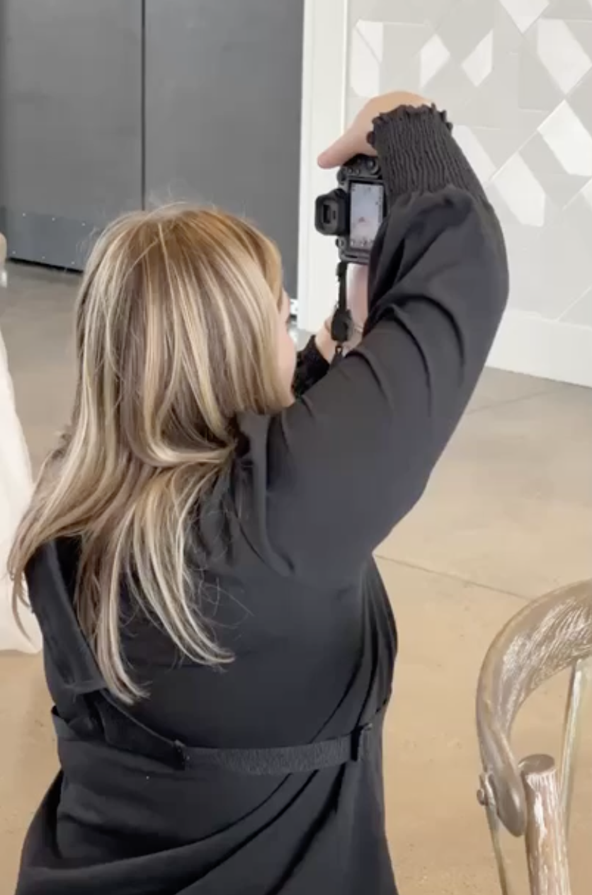 A woman with long blonde hair, wearing a black jacket, is taking a photograph with a camera indoors. Part of a beige or wooden chair is visible in the foreground.