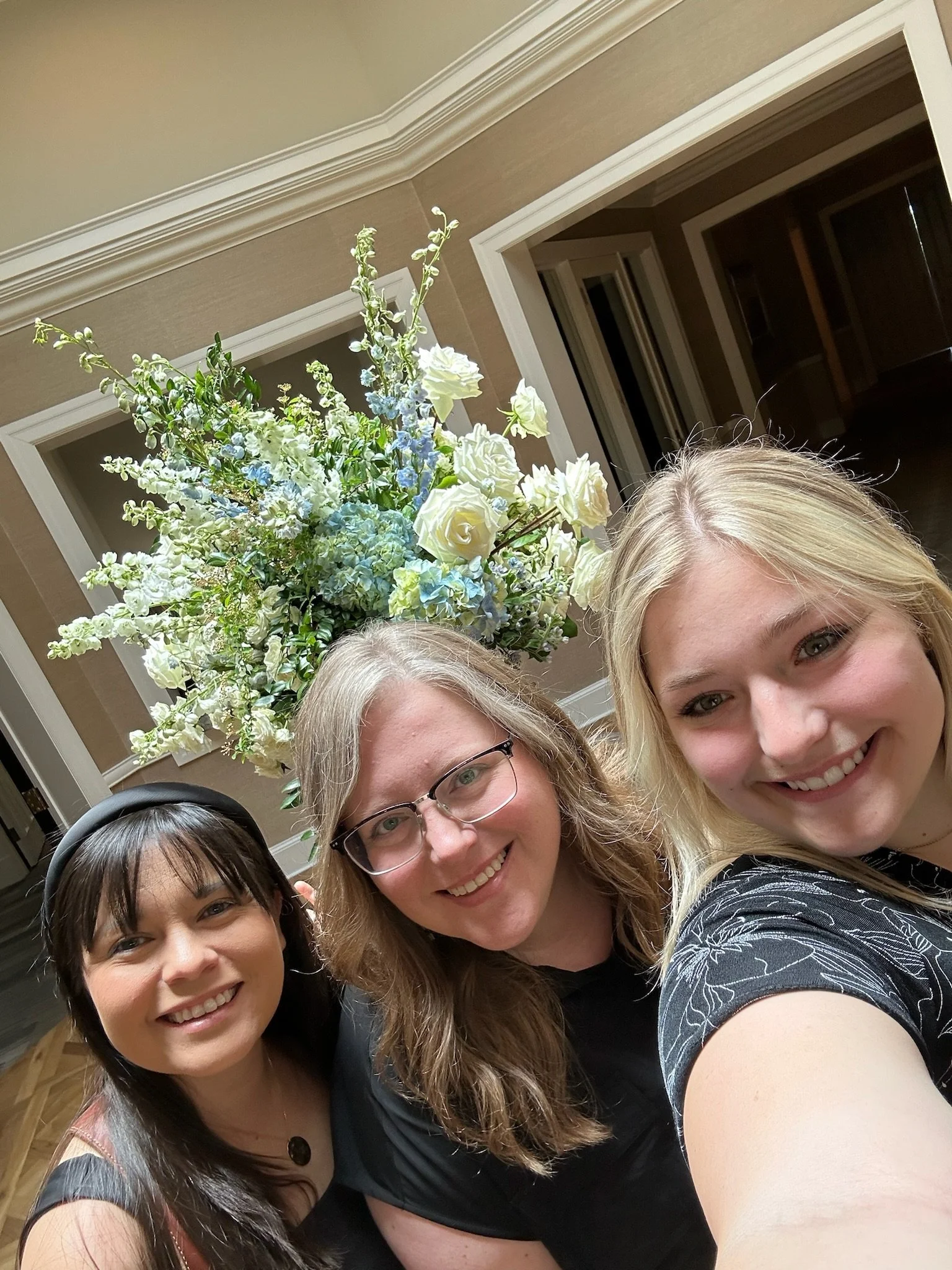 Group of four women, smiling, taking a selfie indoors with a large flower arrangement in the background.