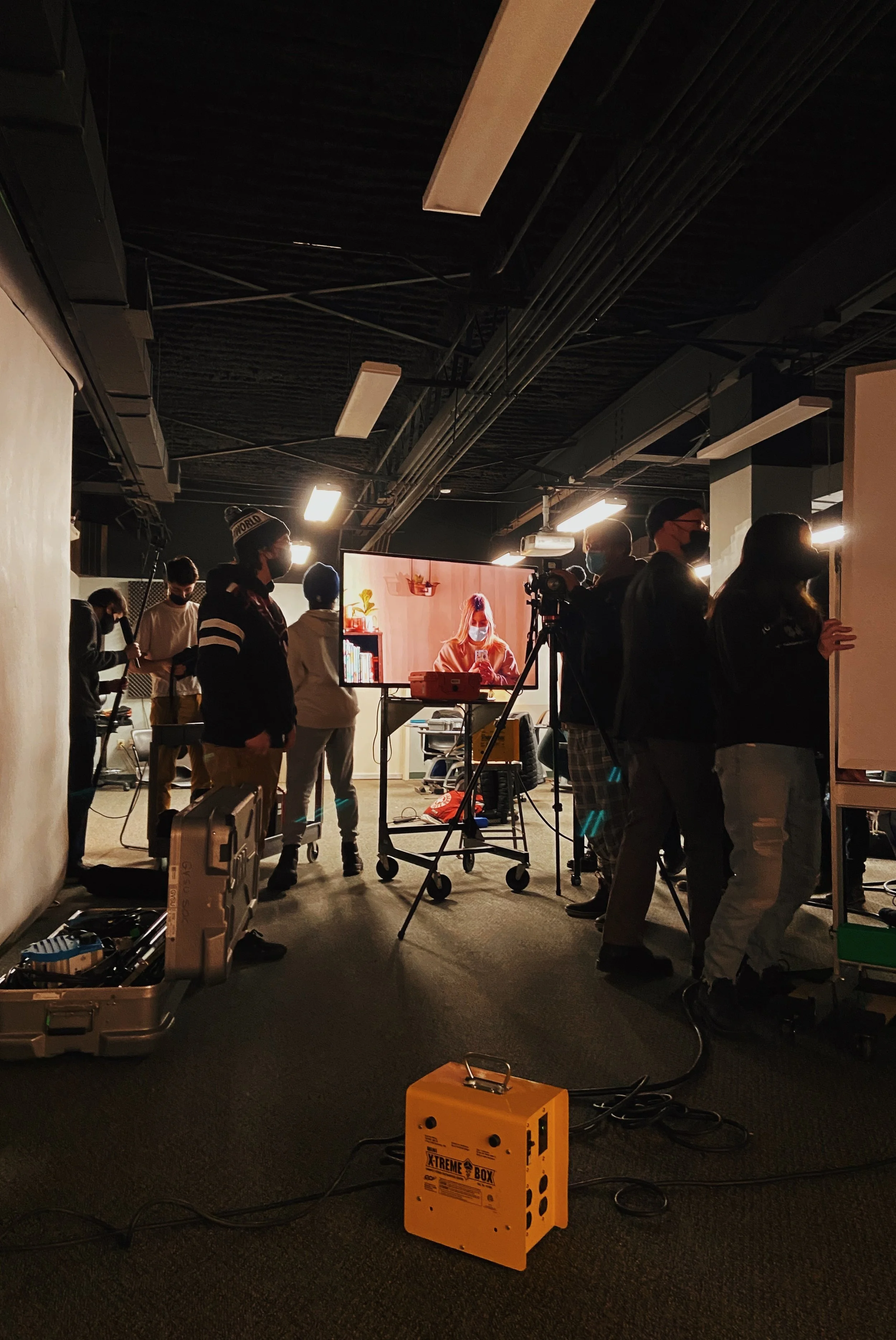 Group of people working in a dimly lit room with equipment, a woman on a monitor, and various tools and cables.