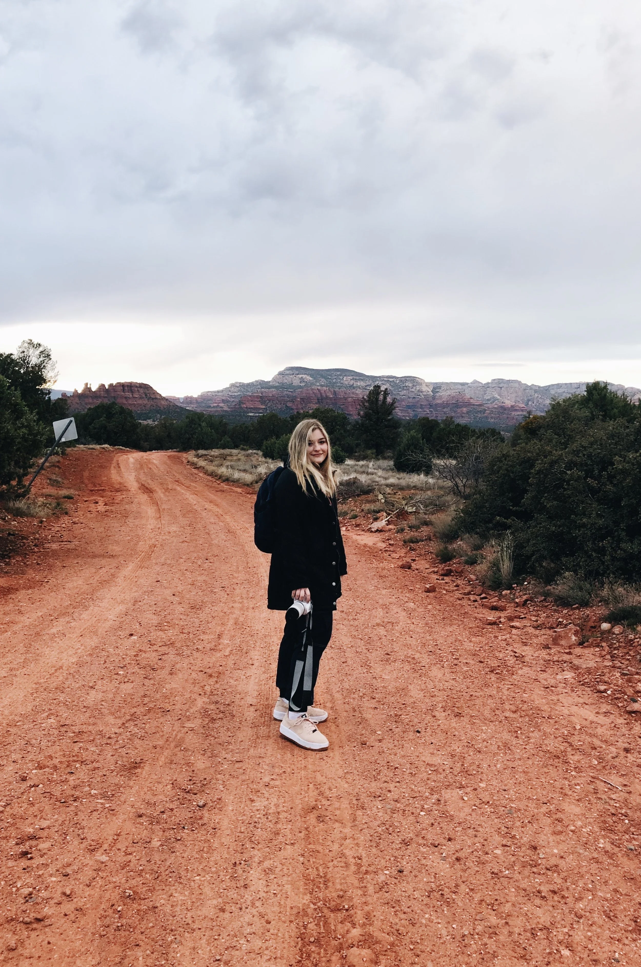 A young woman standing on a dirt road in a desert landscape with red rock formations and trees, holding a camera and wearing sneakers, black pants, and a jacket, under cloudy skies.