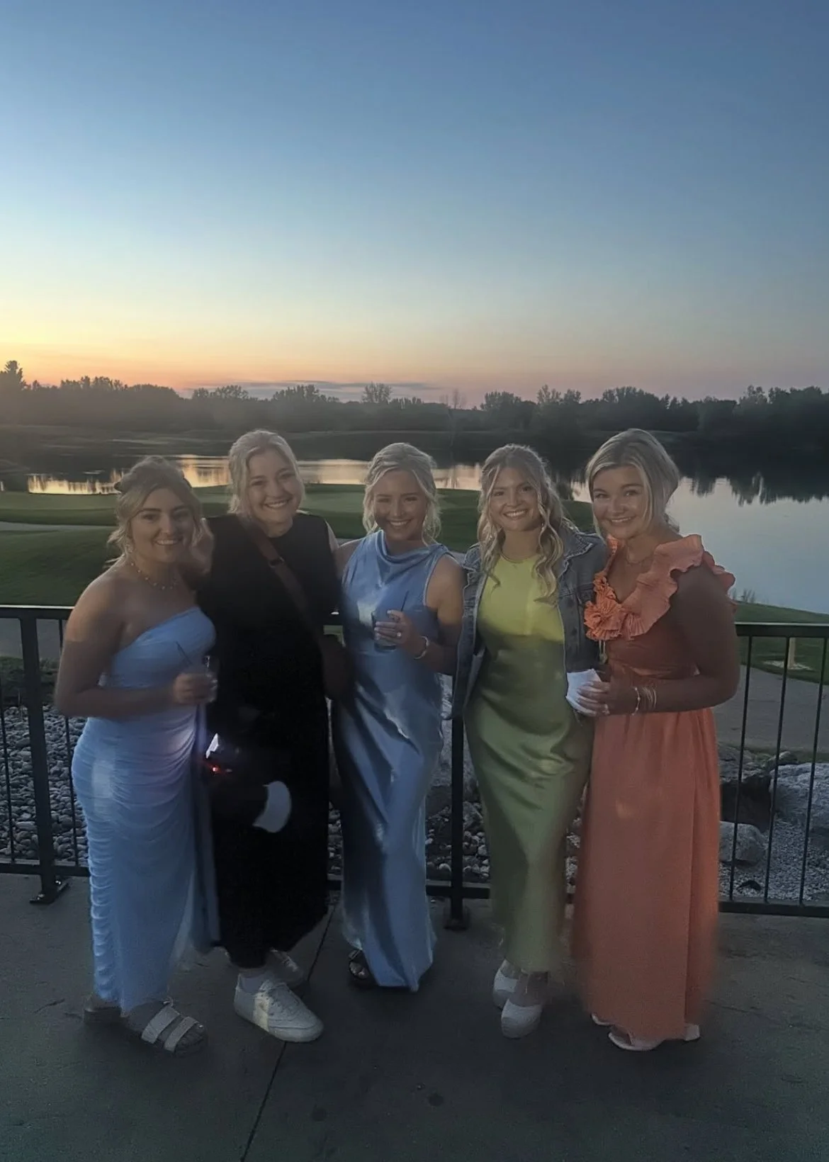 Five women standing together outdoors at sunset, wearing colorful dresses, holding drinks, and smiling at the camera.