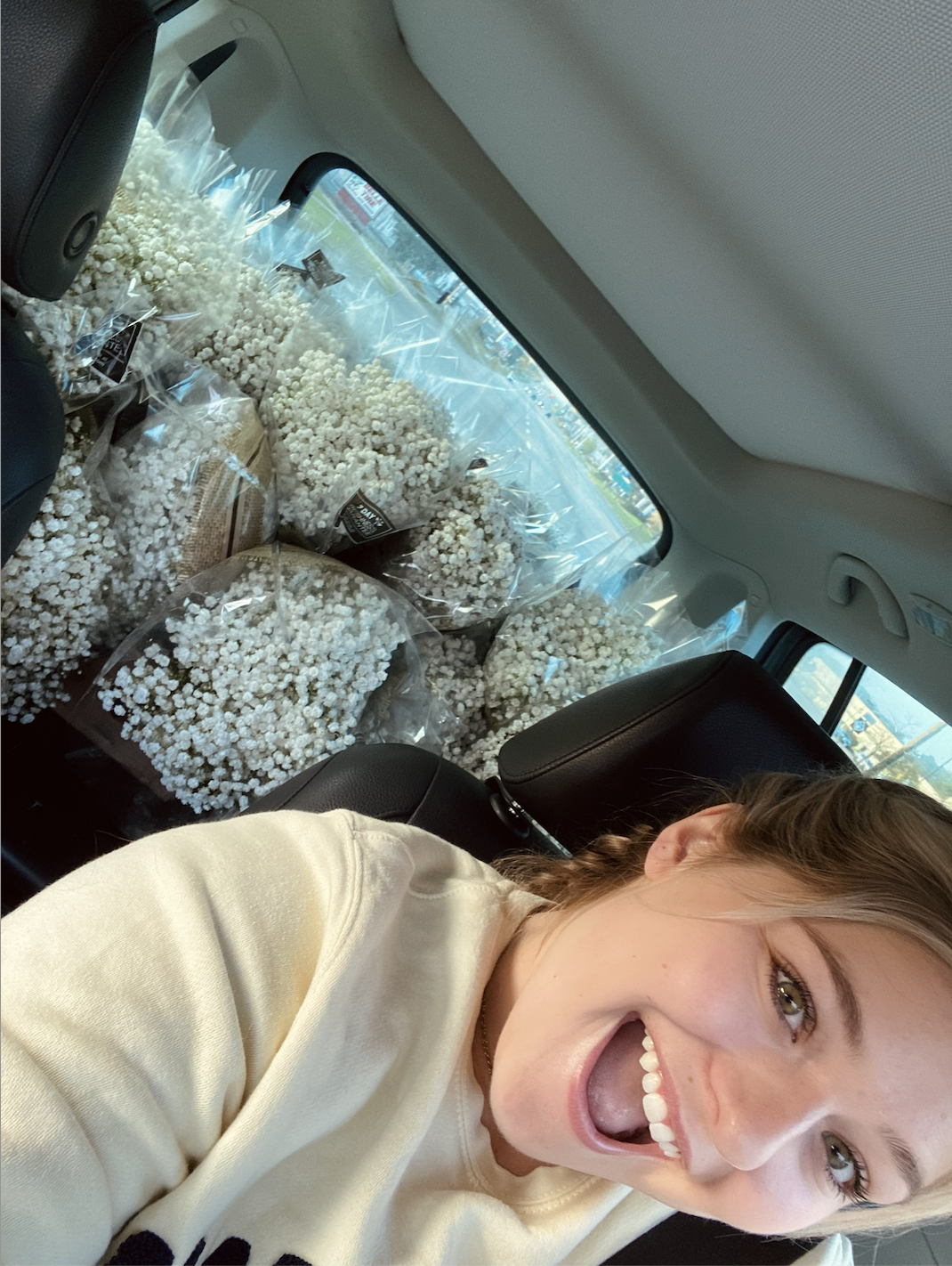 Young woman with brown hair taking a selfie inside a car, with multiple bouquets of white flowers wrapped in plastic on the back seat.