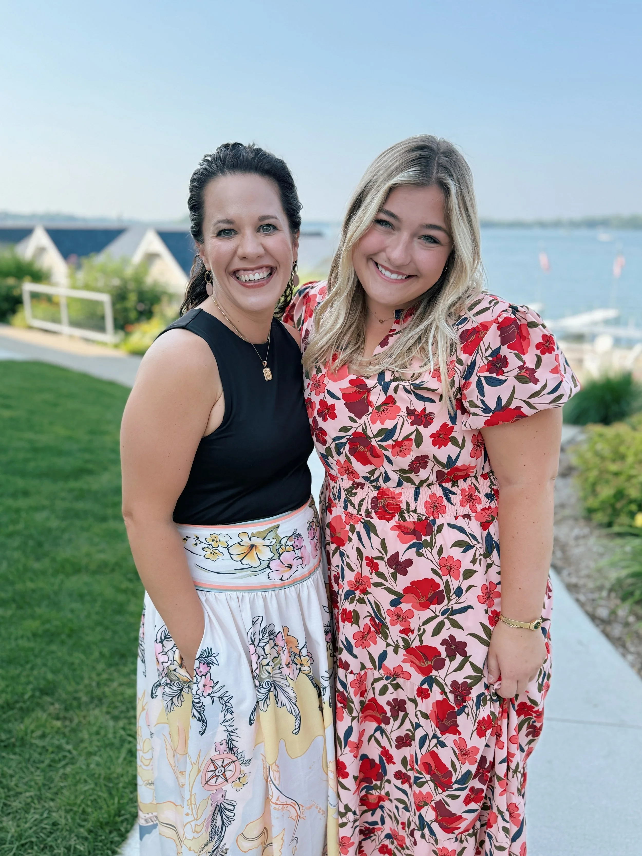 Two women standing outdoors near a body of water, smiling, with houses in the background, dressed in floral dresses.