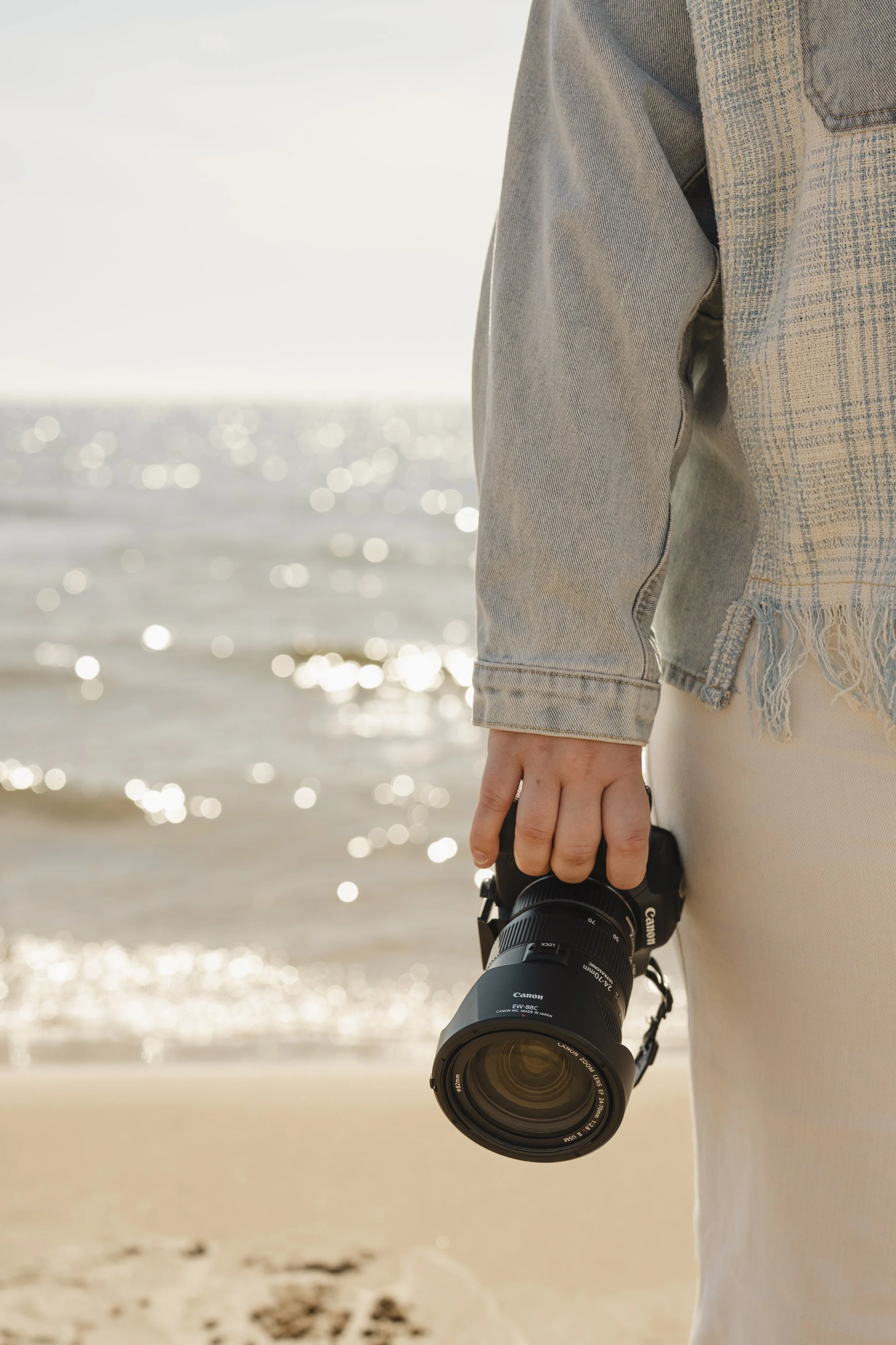 Person wearing a light-colored plaid jacket and jeans holding aCanon camera with a large telephoto lens at a sunny beach.